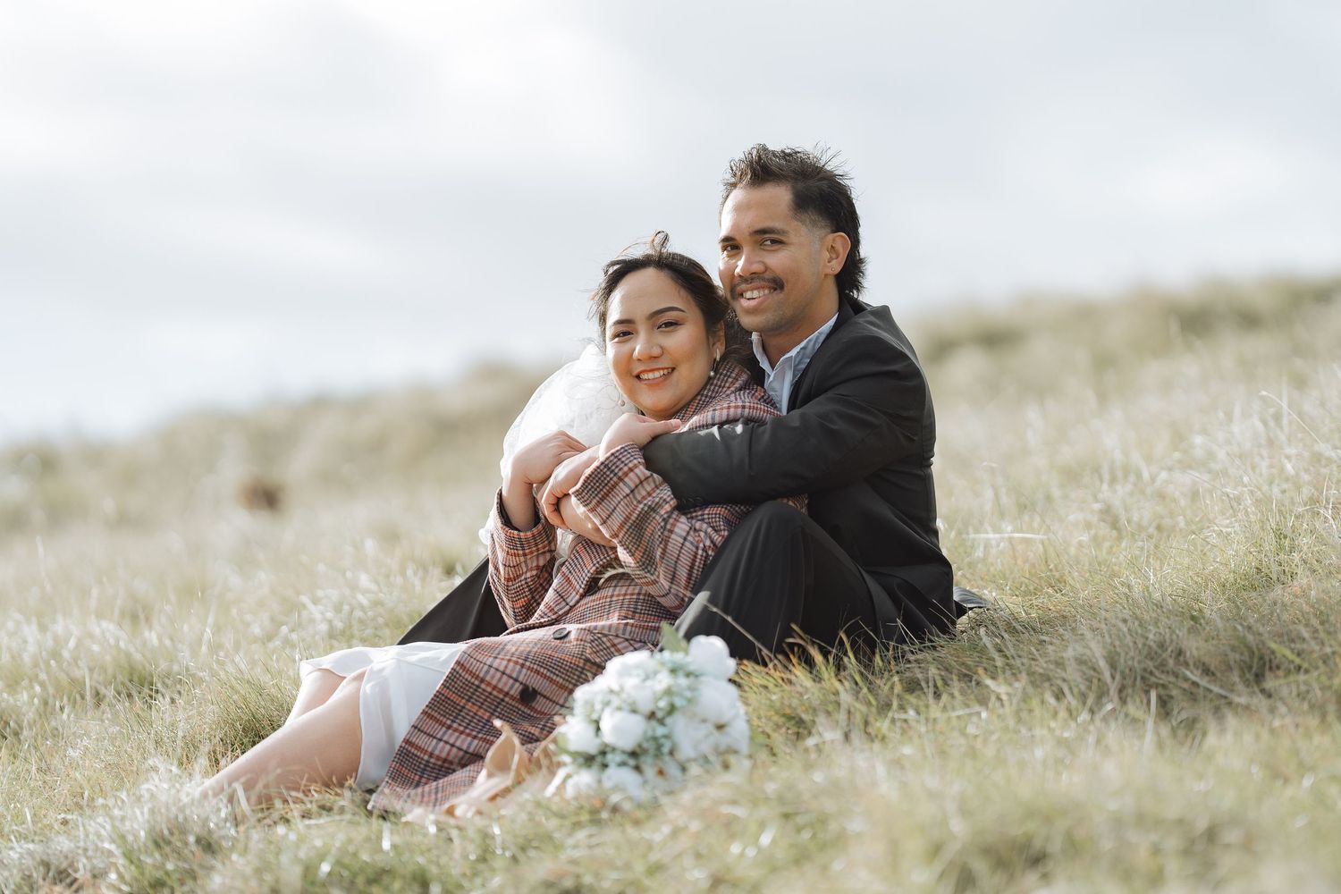 Couple sitting together on grassy hillside under cloudy sky.