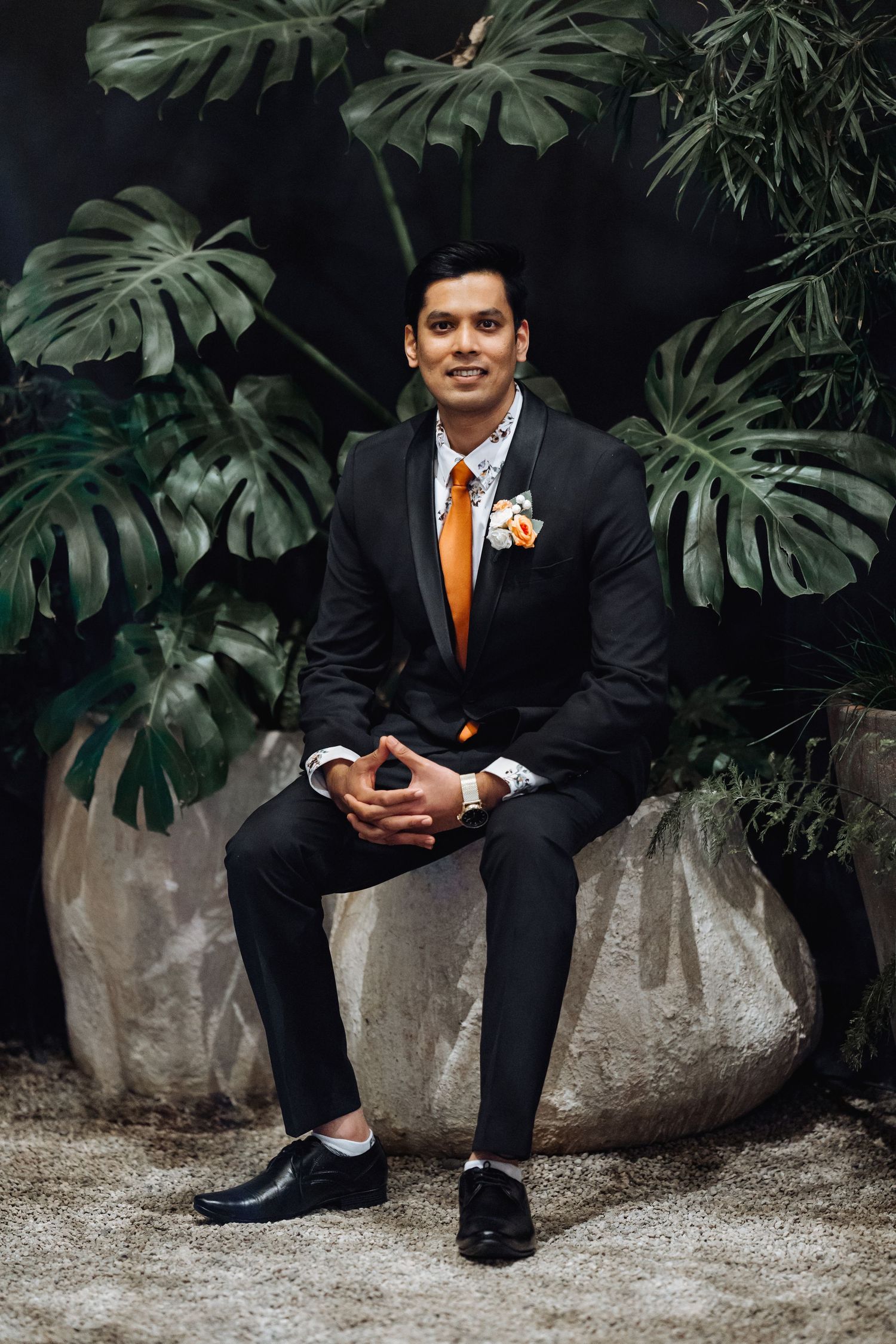 A formal portrait of someone in a black suit with orange tie against large monstera leaves.