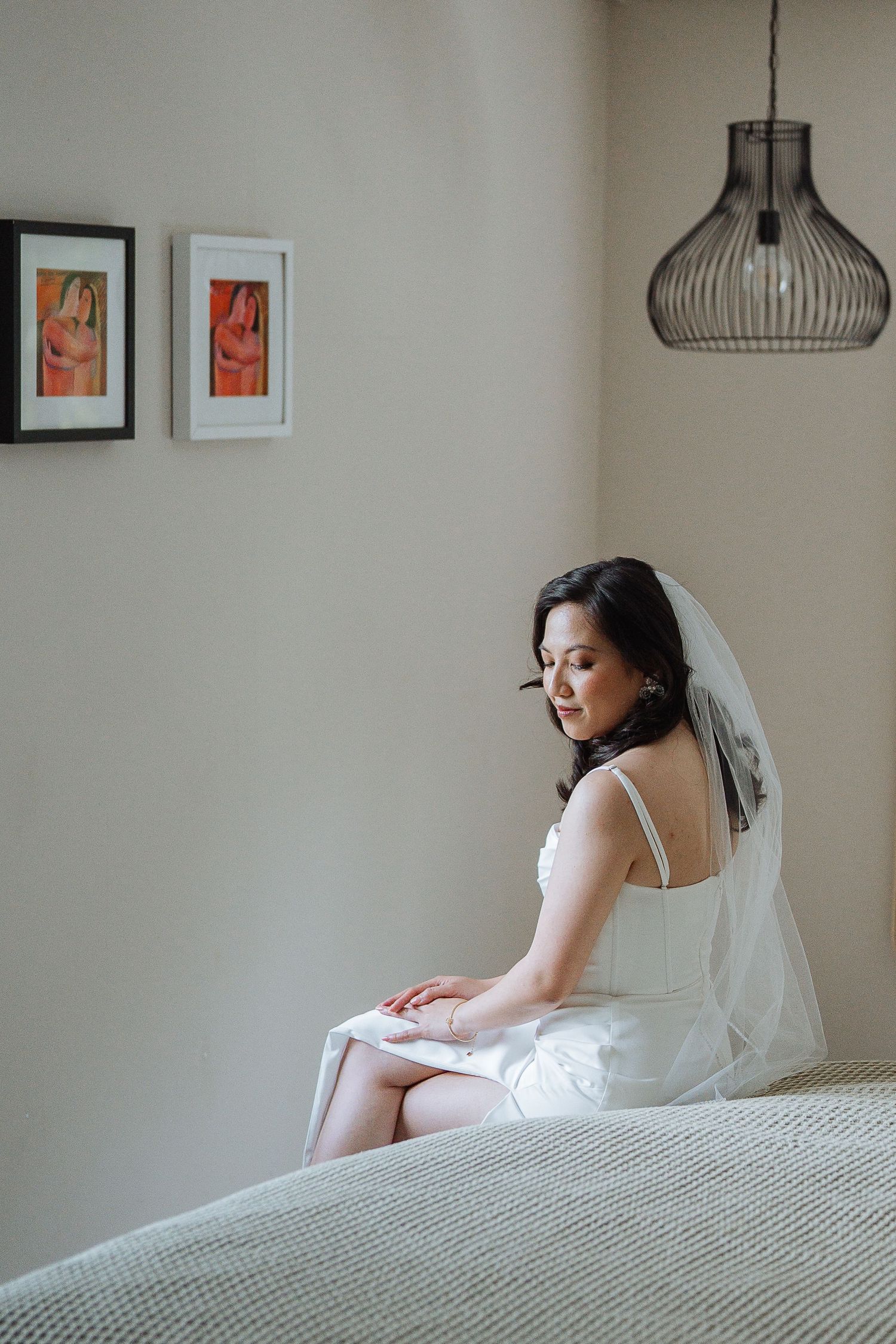 A bride in white dress and veil sits contemplatively on a bed near a pendant light.
