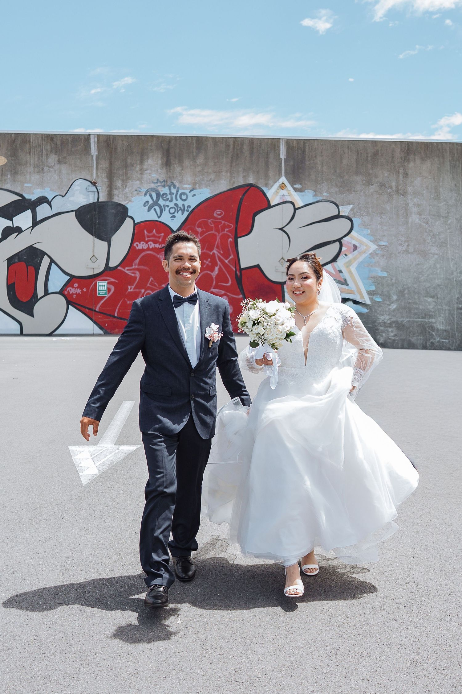 A couple in wedding attire runs playfully along a wall with red graffiti art.