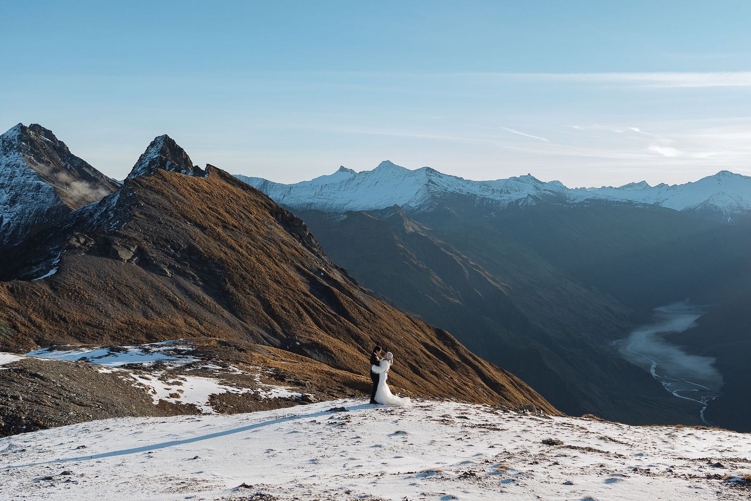 Couple in wedding attire standing on a snowy mountain peak with panoramic alpine views and a winding river below.