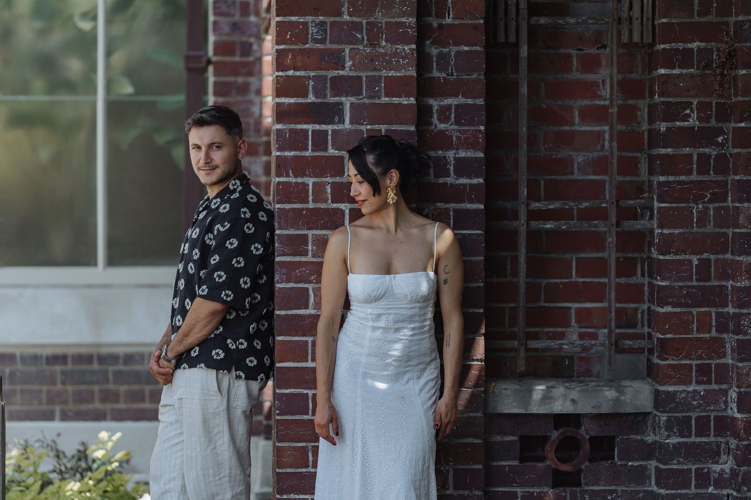 Couple in coordinated outfits posing against brick wall with greenery in an outdoor setting.