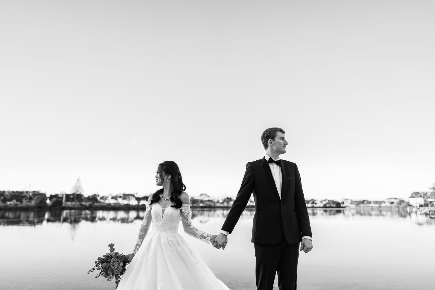 A wedding couple walks along a snowy winter landscape with a lake and trees in the background.