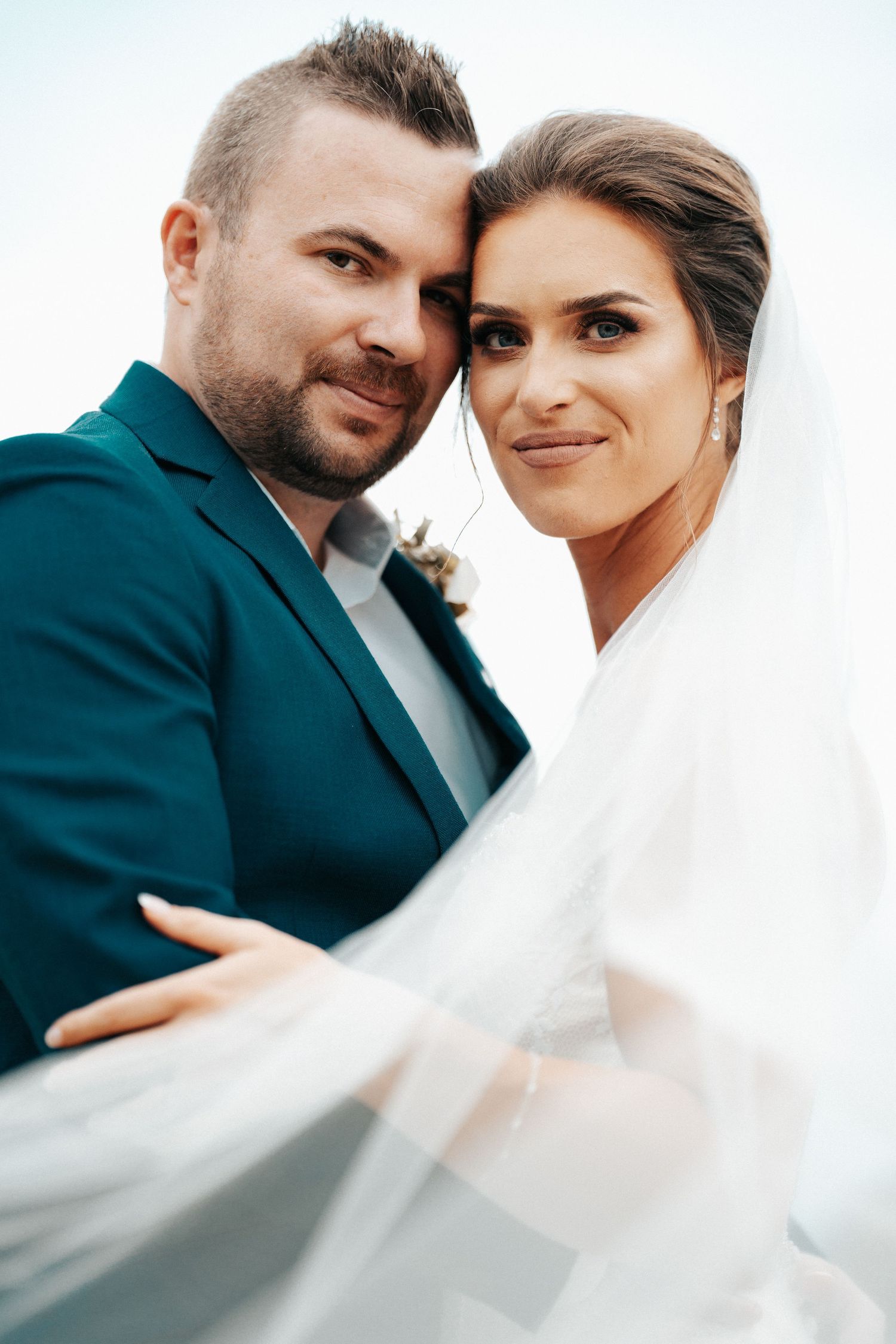 Intimate wedding portrait against bright sky with flowing veil detail.