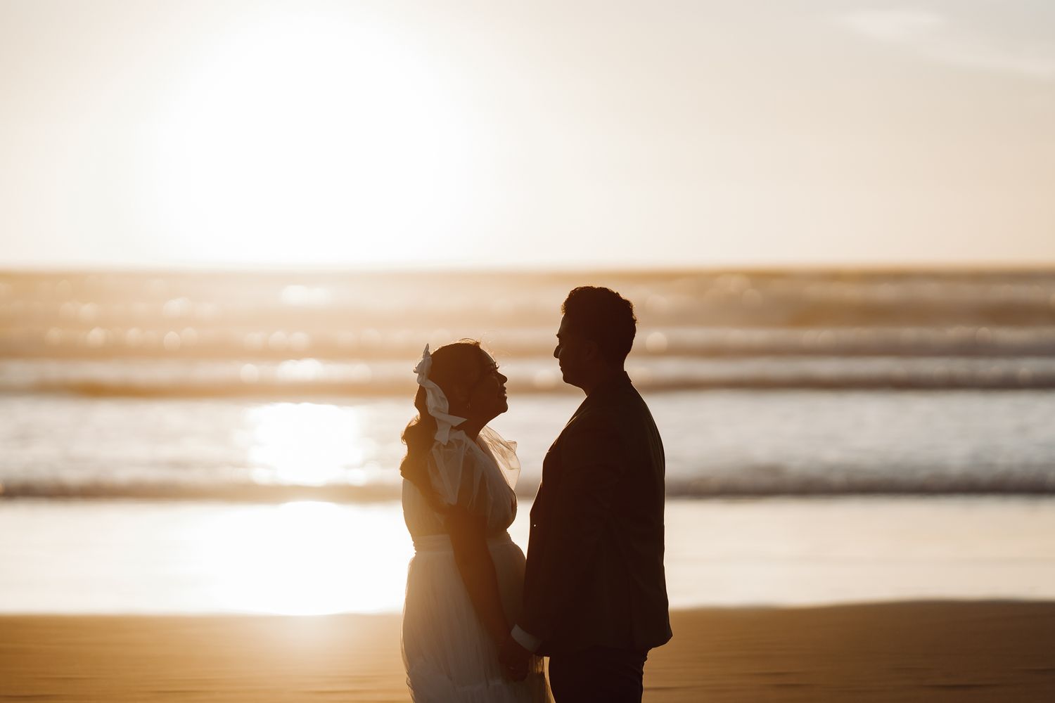 Silhouette of couple standing on beach at sunset with golden light.