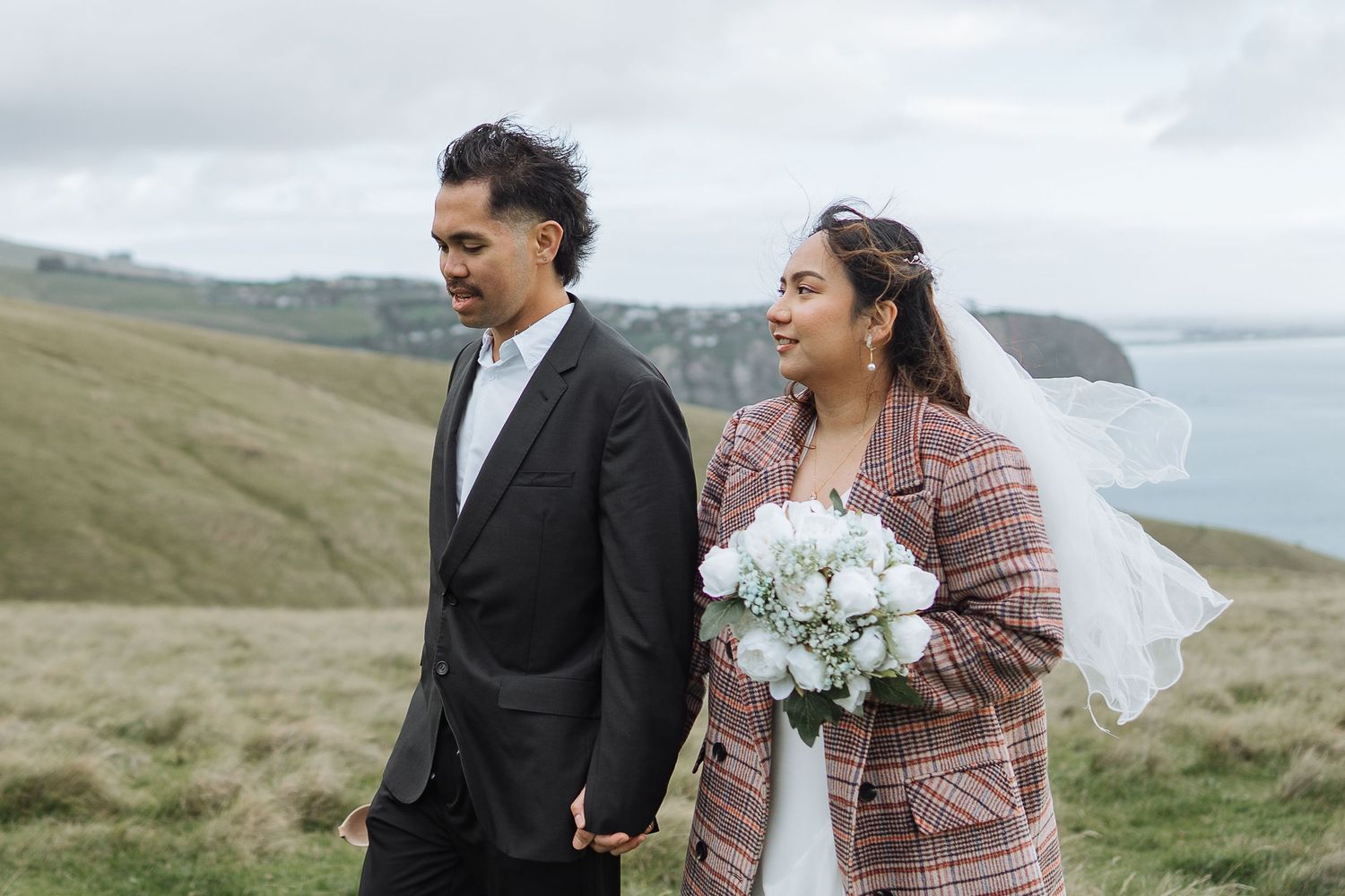 A couple shares a romantic moment on a scenic hillside overlooking the ocean with white flowers.