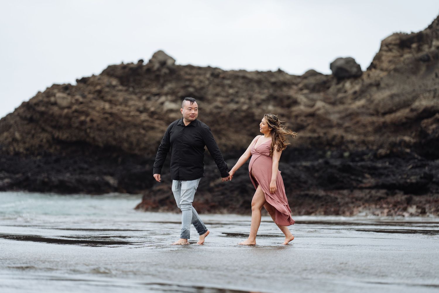 A couple walks hand in hand along a scenic beach with rocky cliffs.