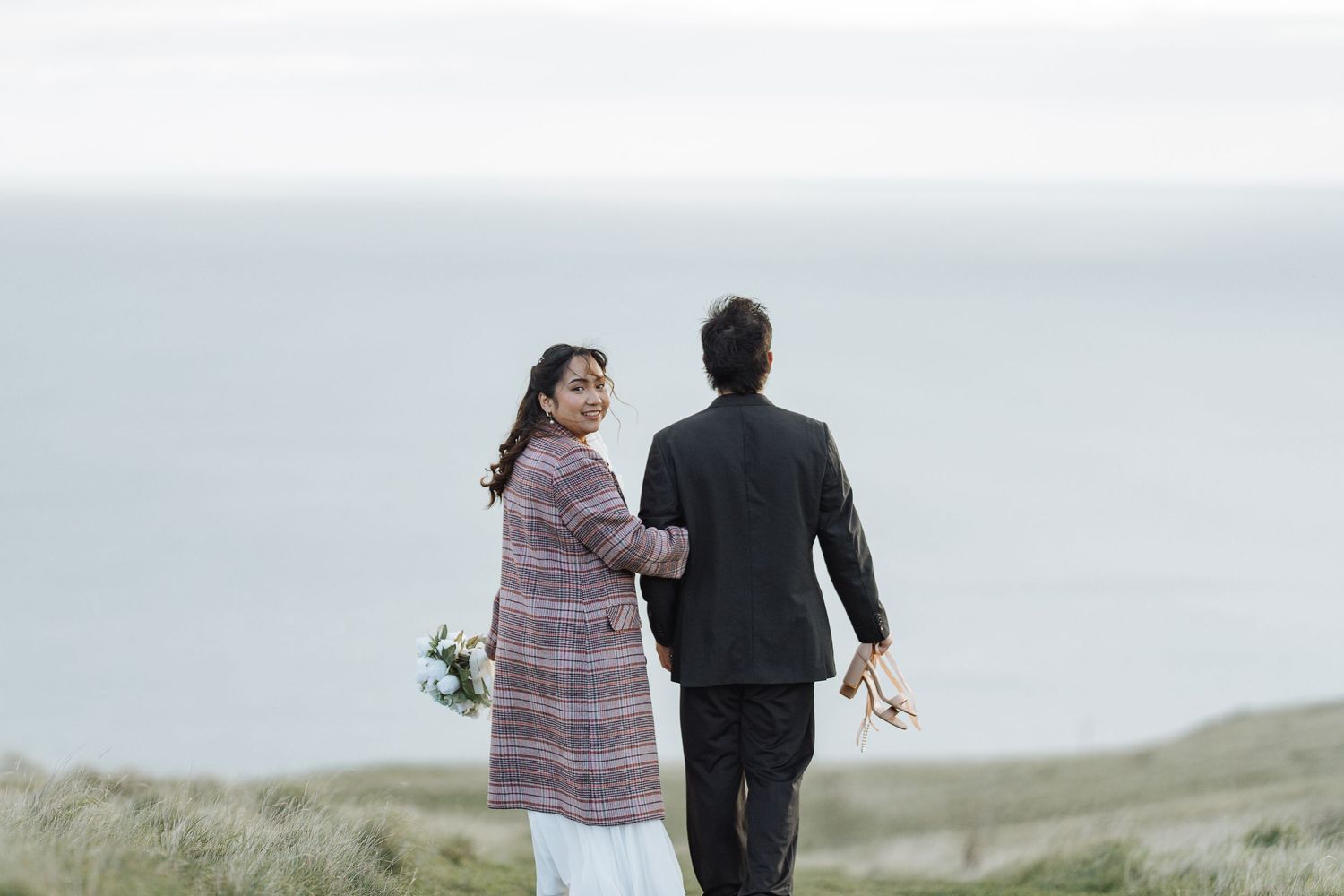 A couple walks hand in hand through tall grass on a coastal hillside.