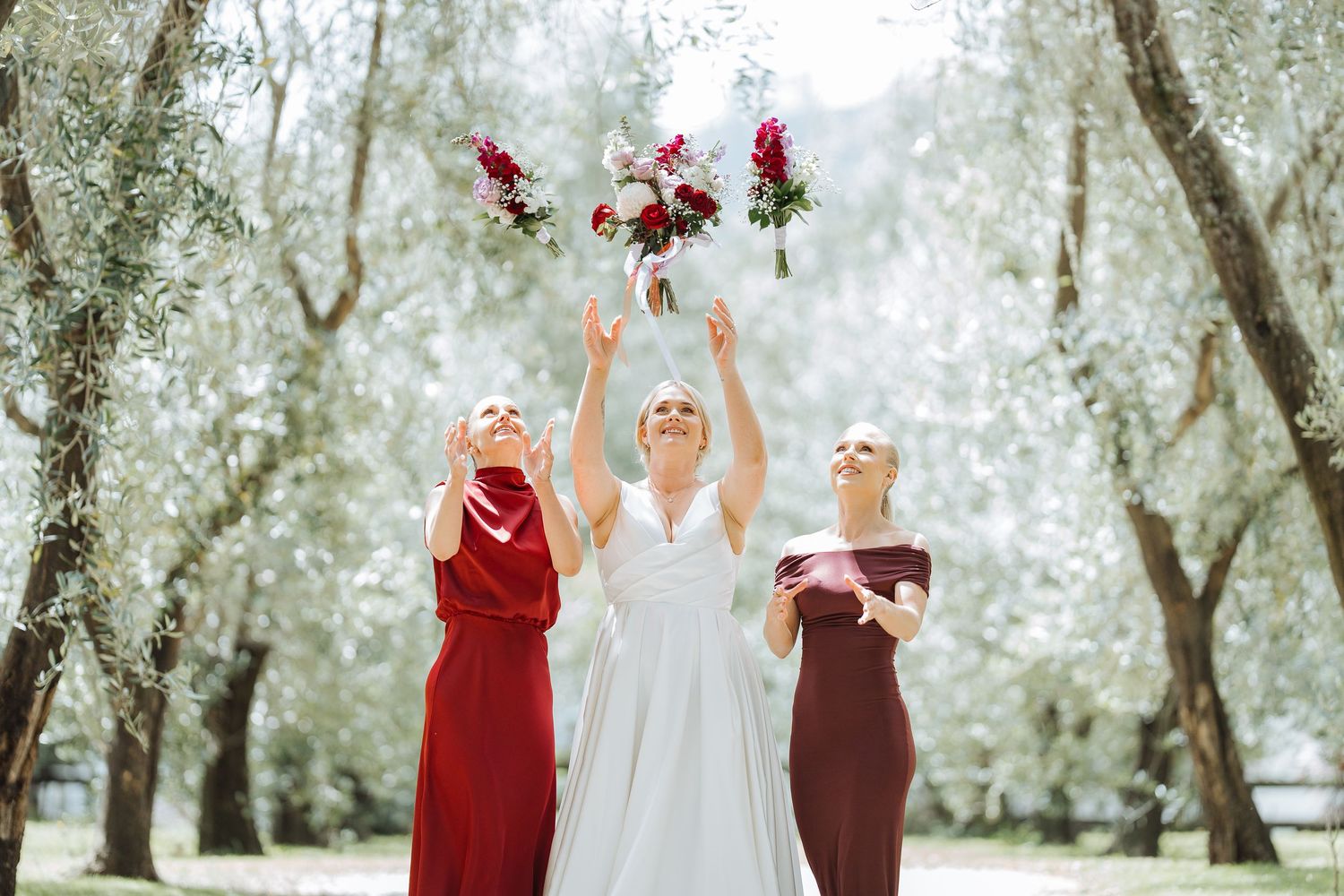 Three people in red and white dresses toss bouquets in a joyful garden celebration.