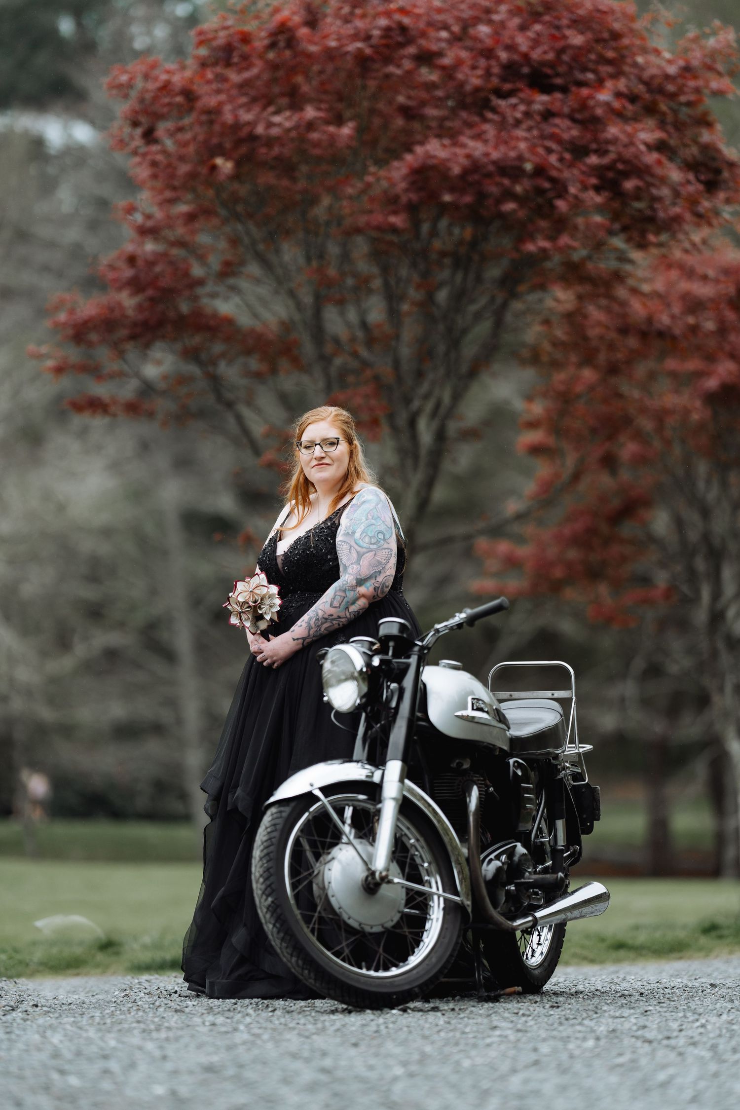 A vintage motorcycle sits against a backdrop of red autumn trees in a moody outdoor photoshoot.