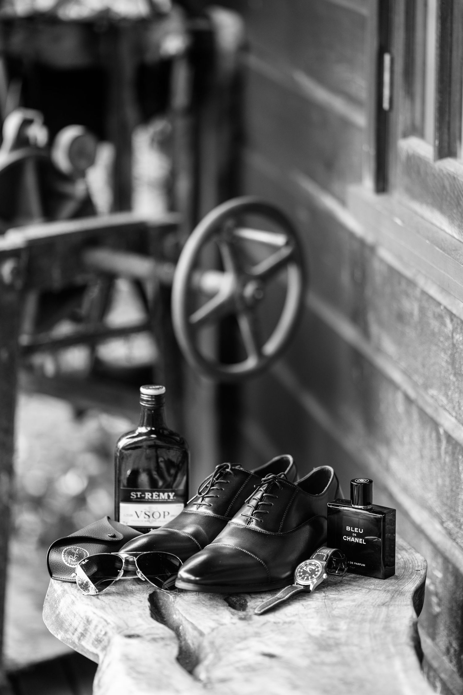 Black and white still life of groom's accessories including shoes, cologne and sunglasses.