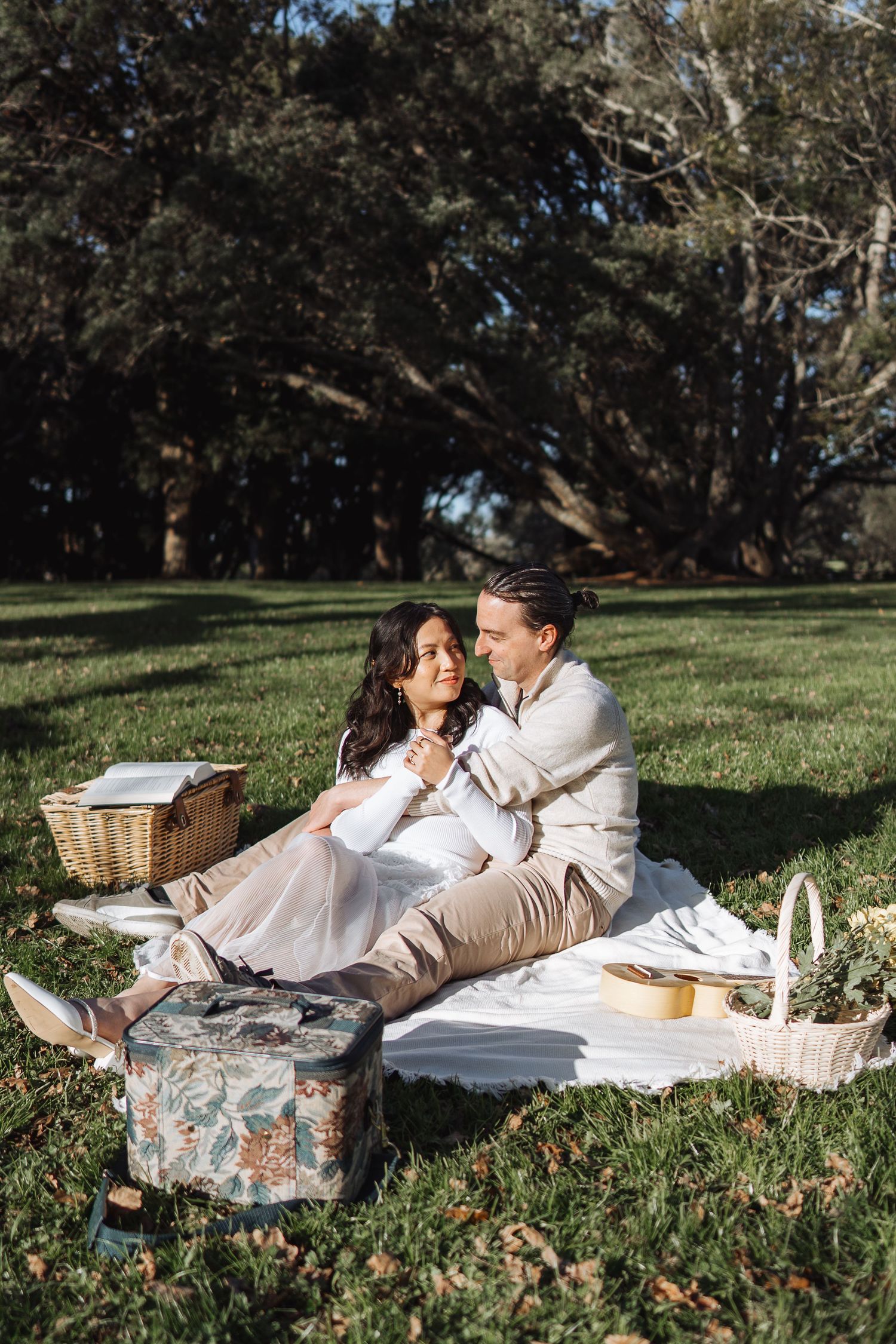 A couple enjoys a romantic picnic on a blanket spread out on grassy ground surrounded by trees.