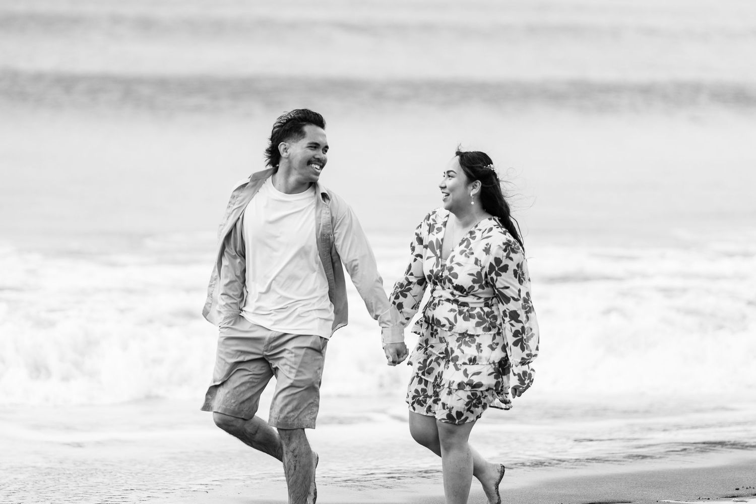 A couple runs playfully along a sandy beach with waves in the background.