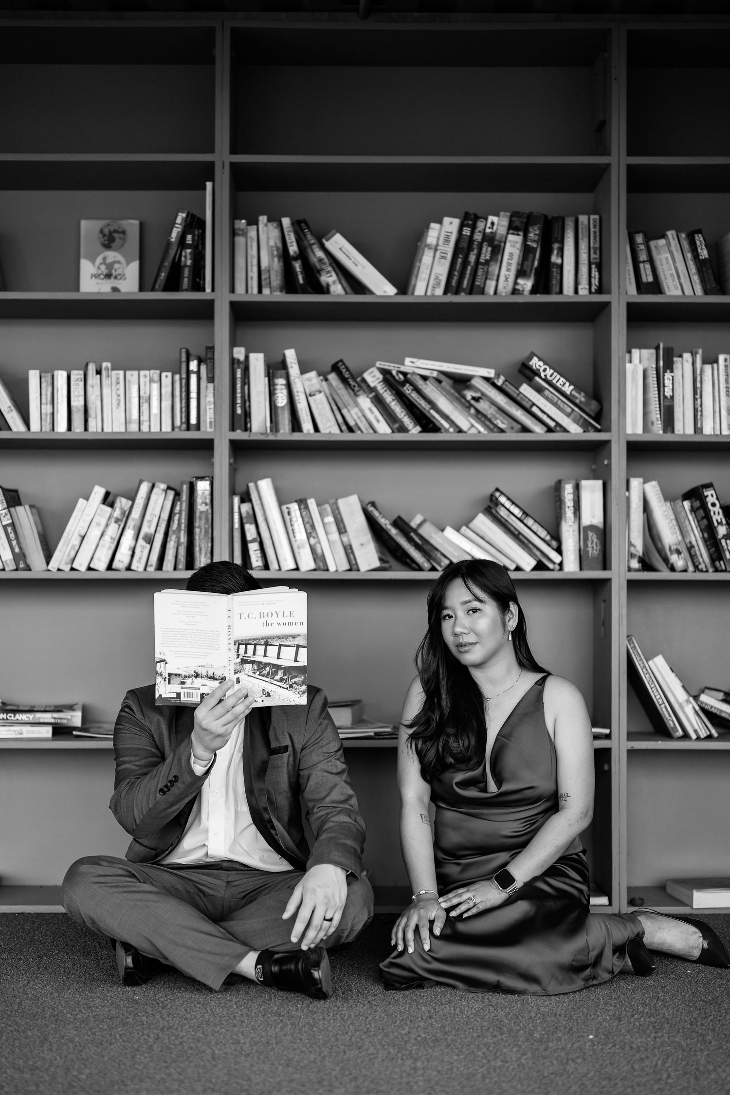 Black and white photo of bookshelves with people sitting on floor in library.