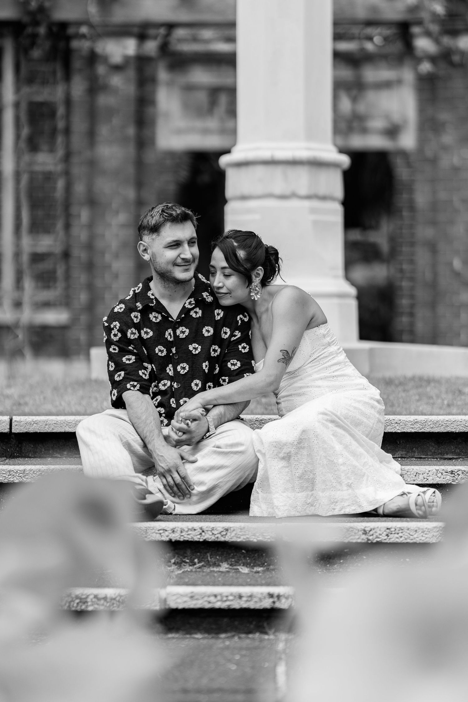 A romantic couple in black and white share an intimate moment on a bench in a garden setting.
