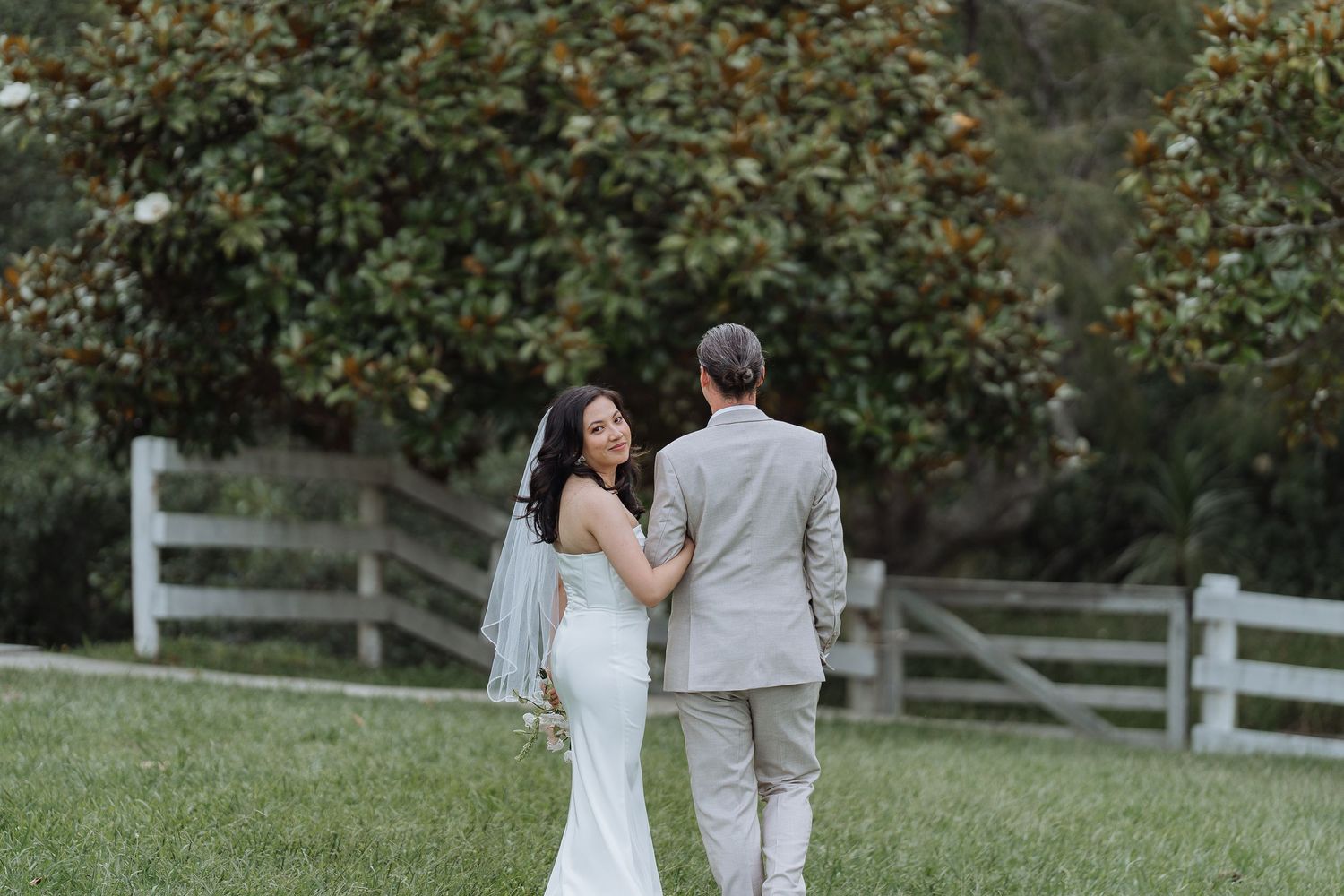 Couple walks together on grassy lawn with rustic fence in background during wedding photoshoot.