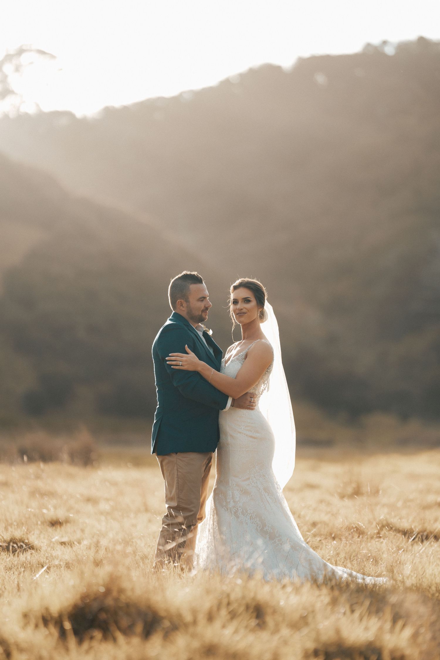 Romantic sunset wedding portrait in golden field with mountains in background.