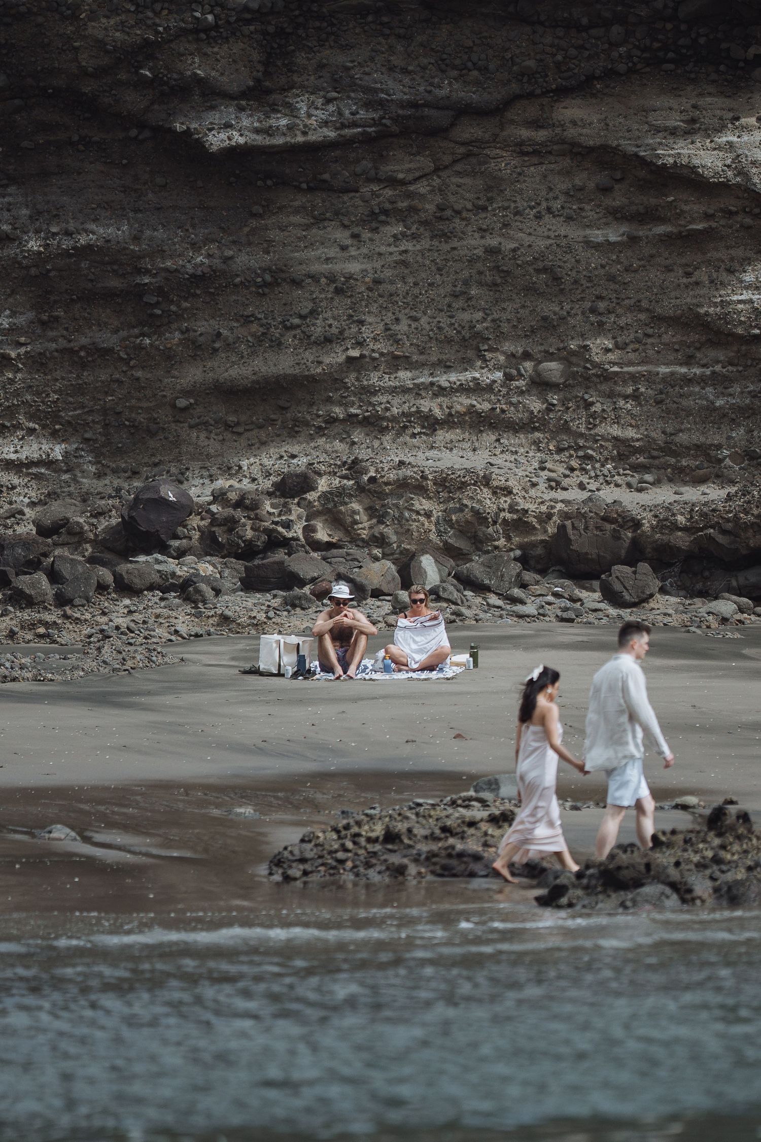 Distant view of couple walking along rocky beach with cliffs in background.