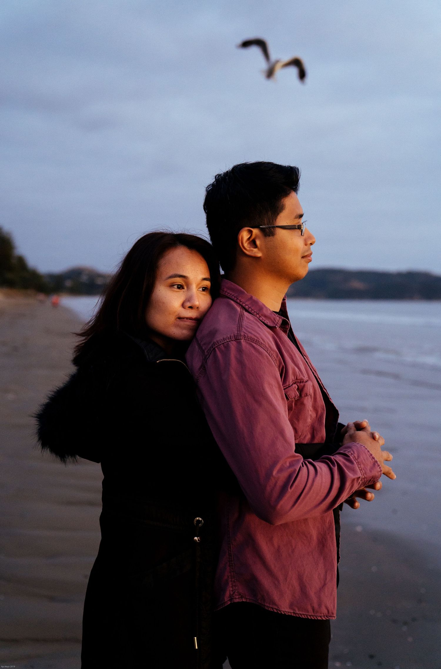 Couple in casual attire standing on beach during sunset with seabirds flying overhead.