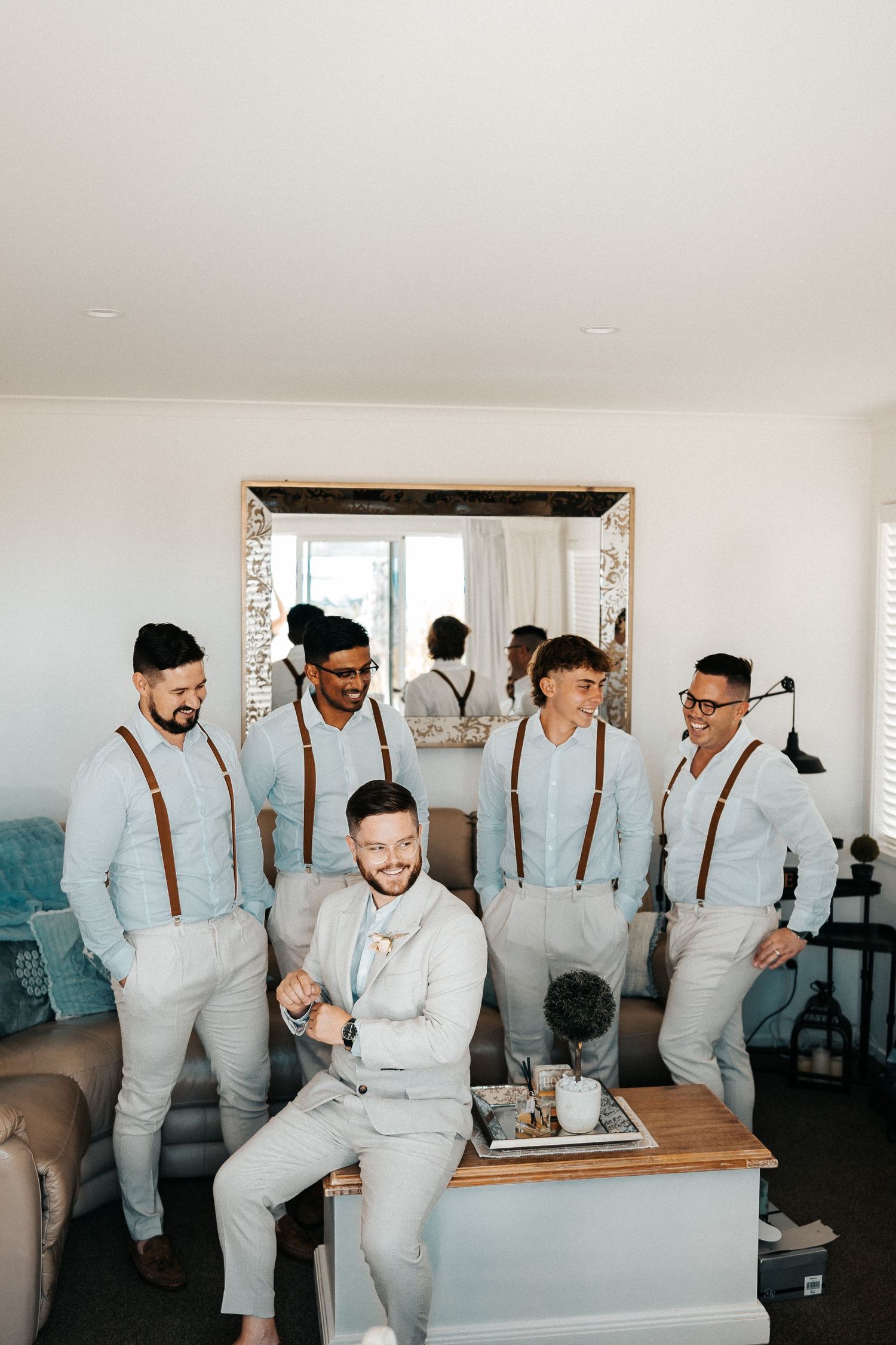 Groomsmen in light gray suits and suspenders gather in a bright indoor space.