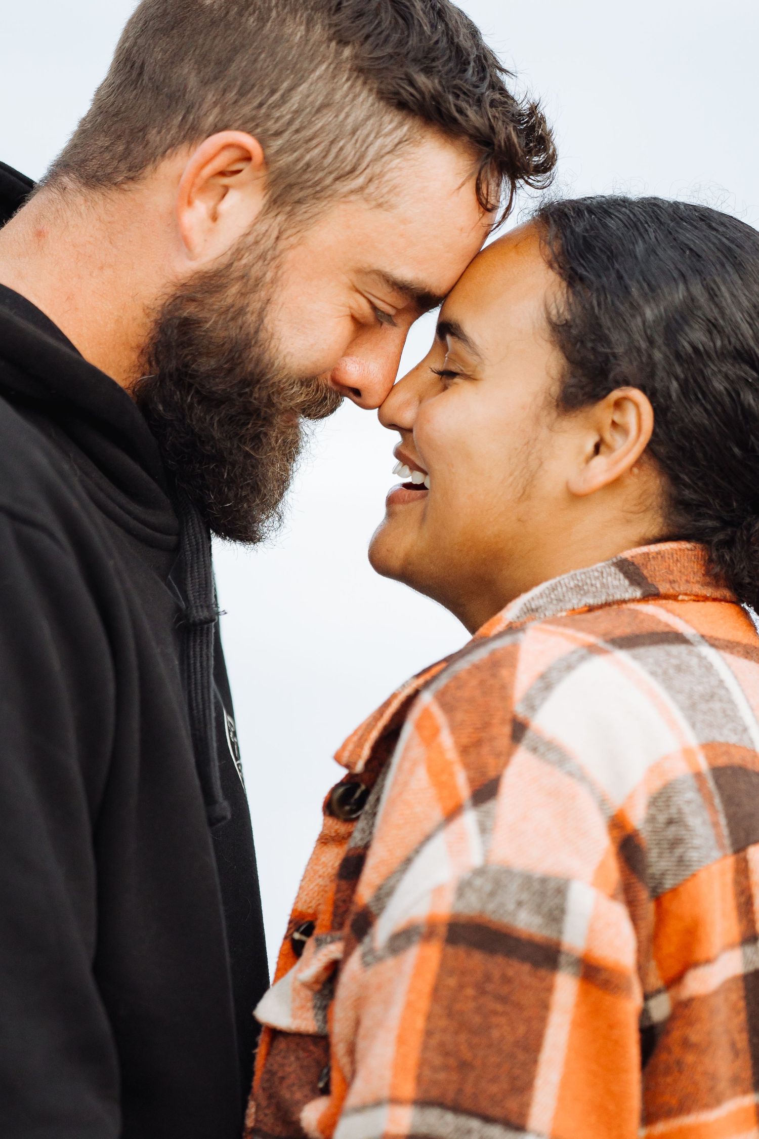 Couple sharing a tender nose-to-nose moment, man with beard in black jacket, woman in orange plaid shirt, smiling.