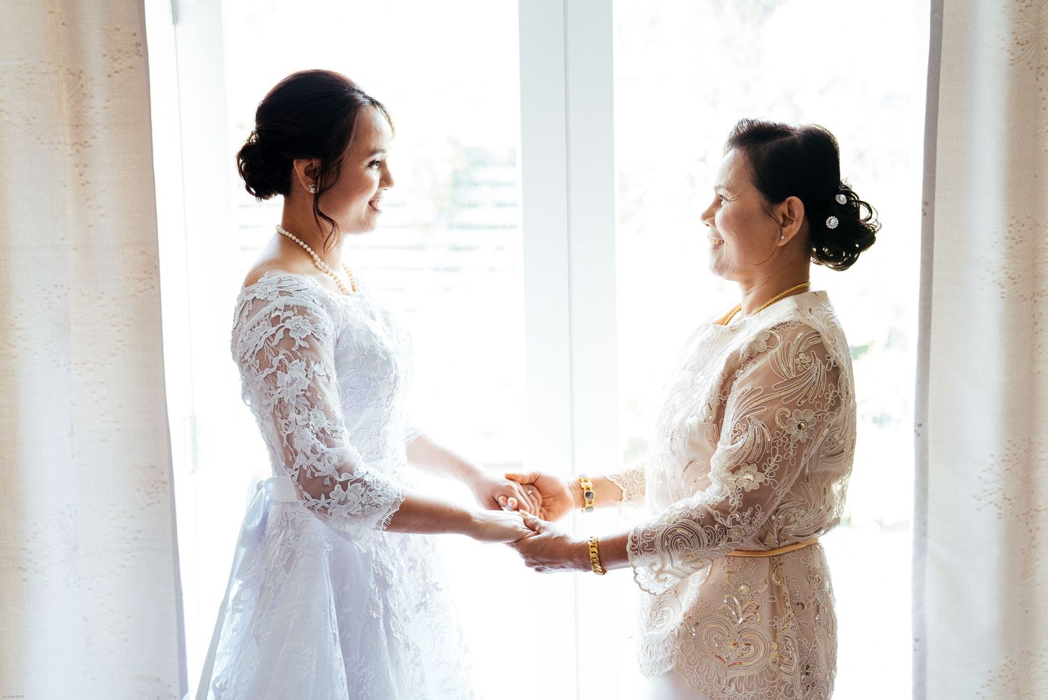 Tender moment captured by window light in elegant white lace dresses.