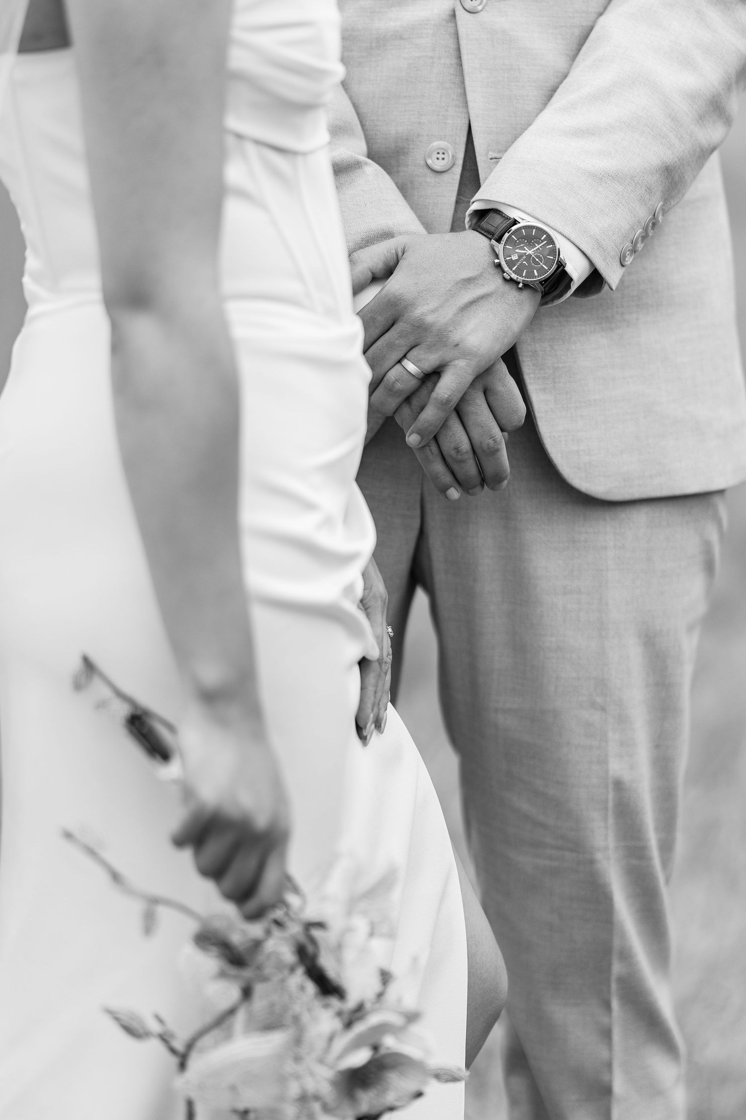 Black and white detail shot of couple holding hands, showing wedding attire and wristwatch.