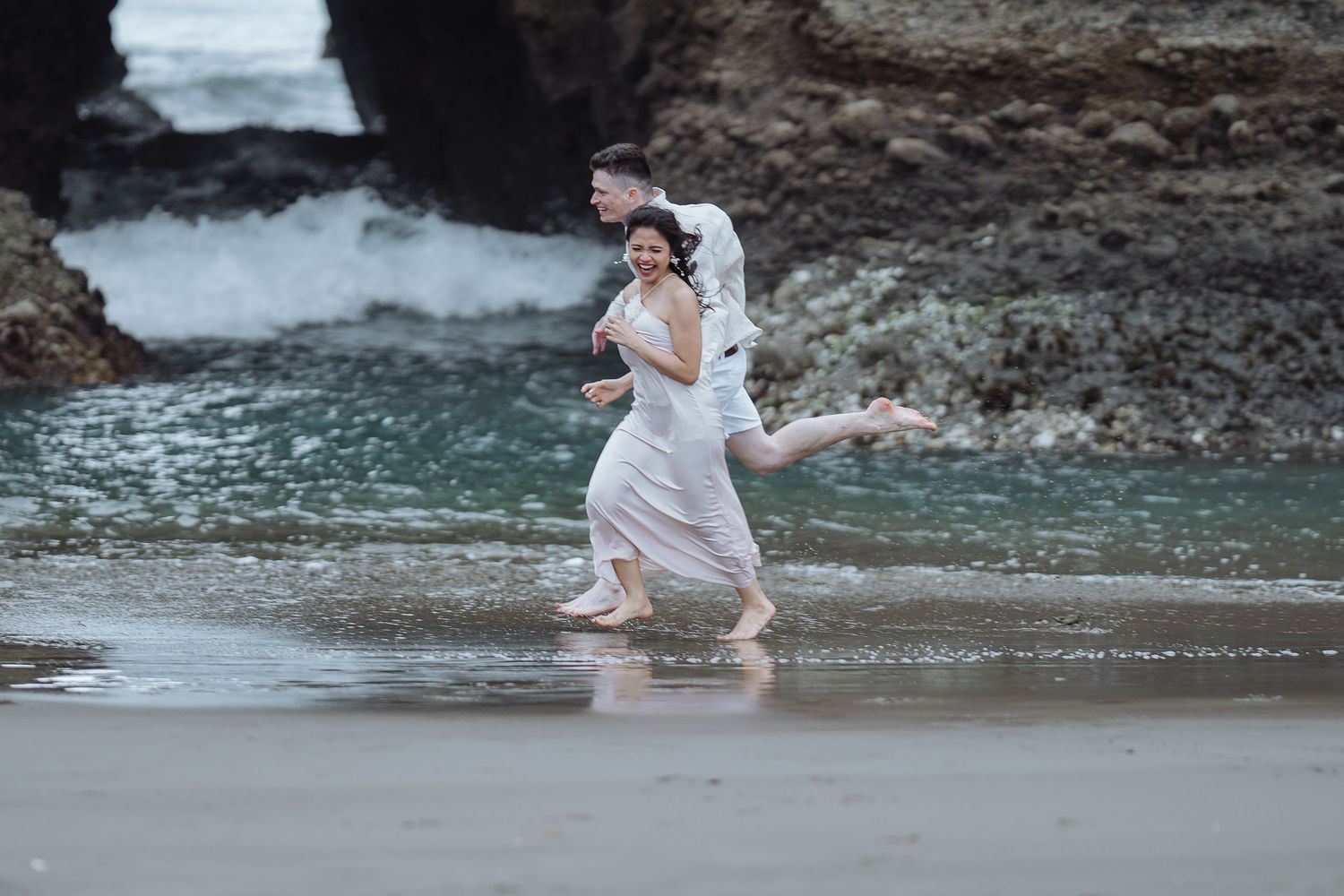 A couple splashes playfully in the ocean water near rocky cliffs during a romantic beach photoshoot.