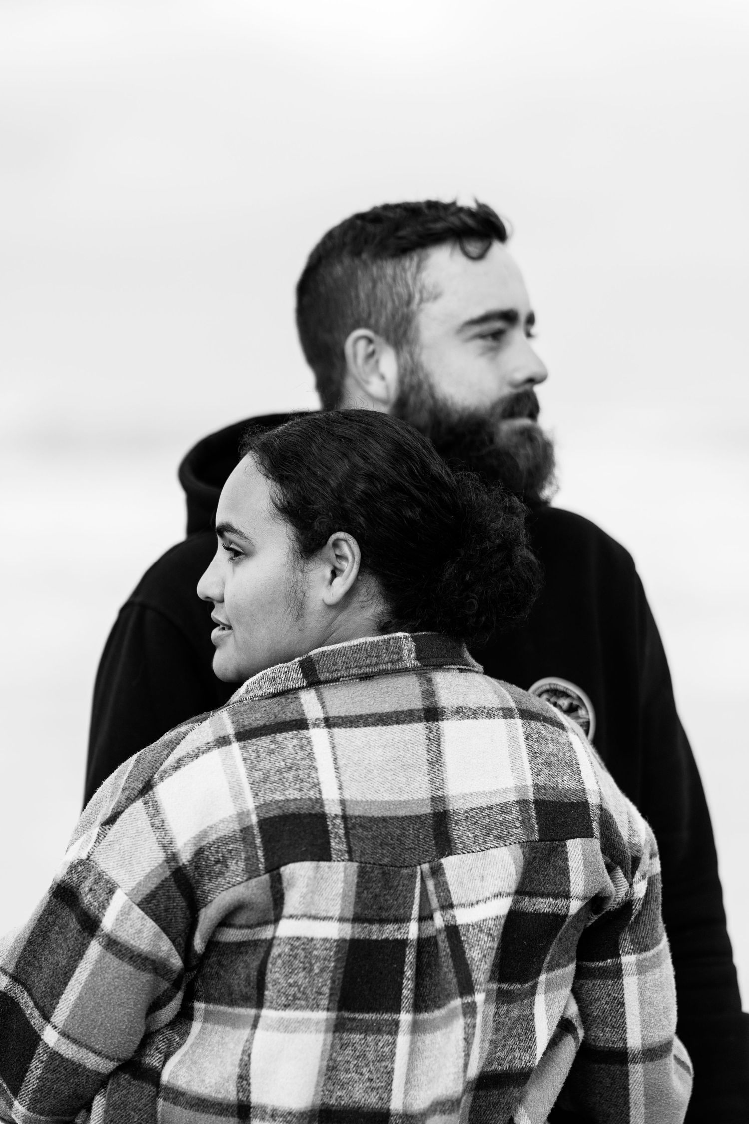 Black and white photo of a bearded man and curly-haired woman in plaid shirt standing together outdoors.