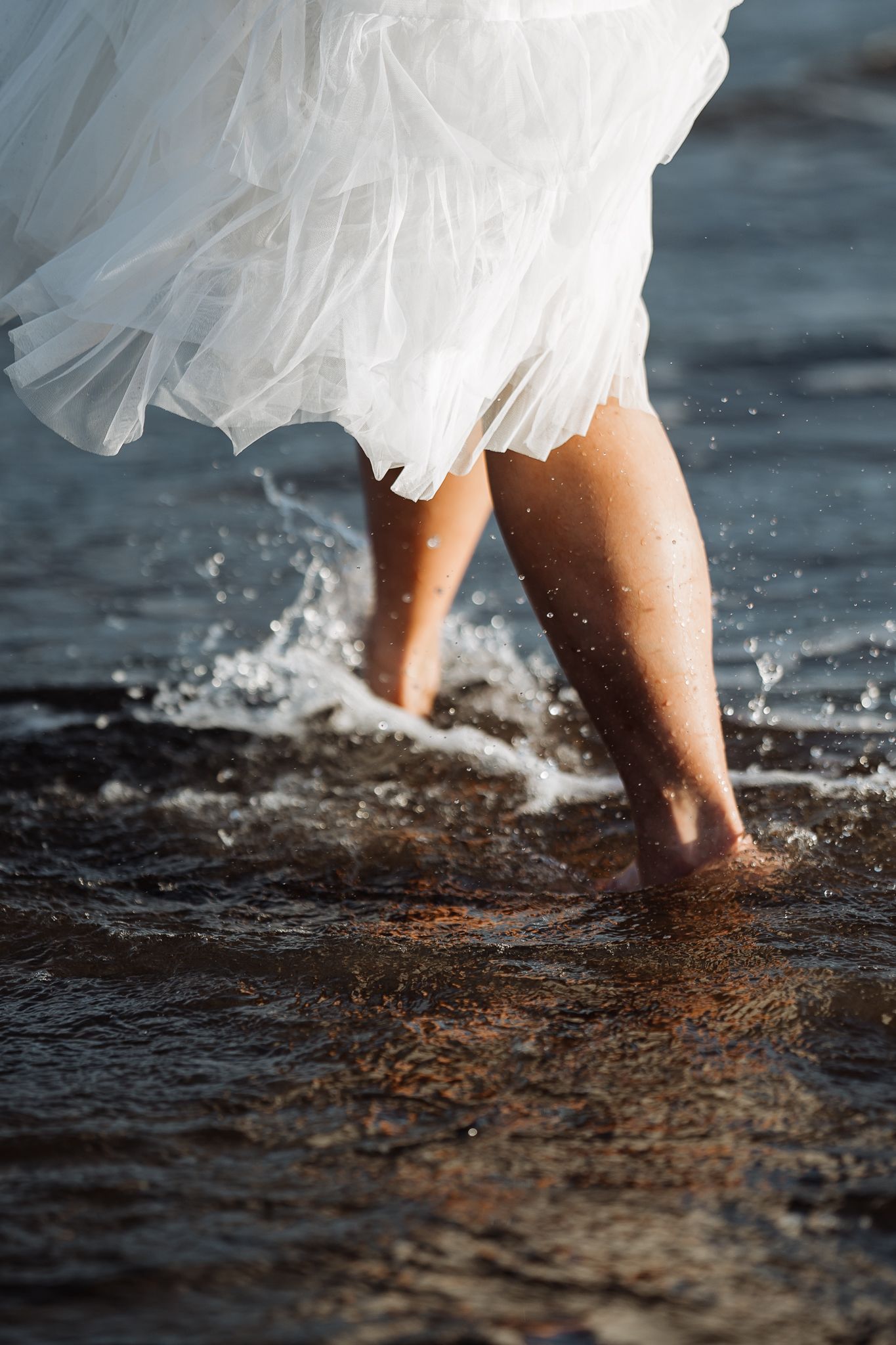 Close-up of bride's legs splashing in ocean water while wearing flowing white dress.