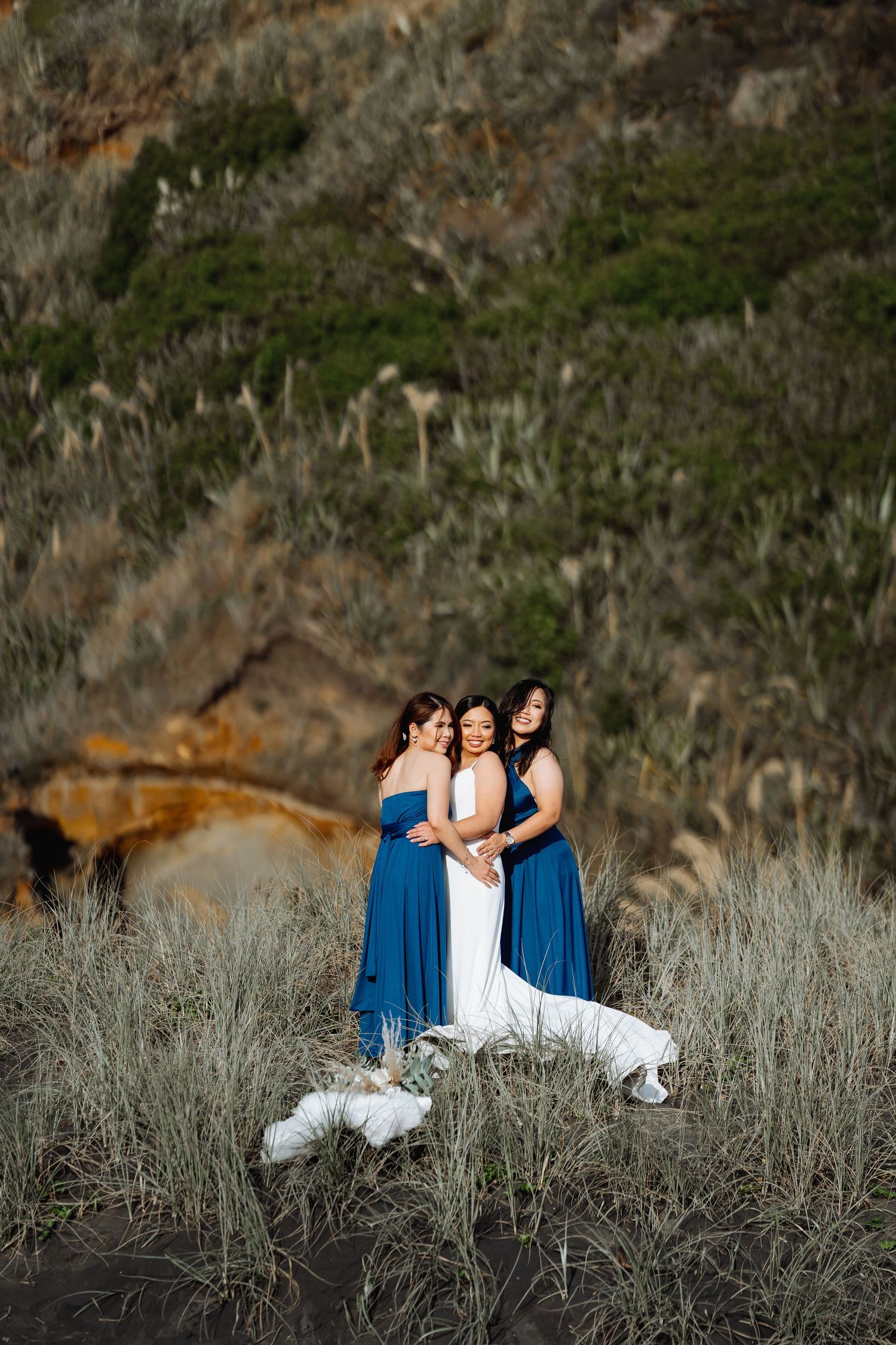Three women in blue and white dresses pose together on a grassy hillside at sunset.