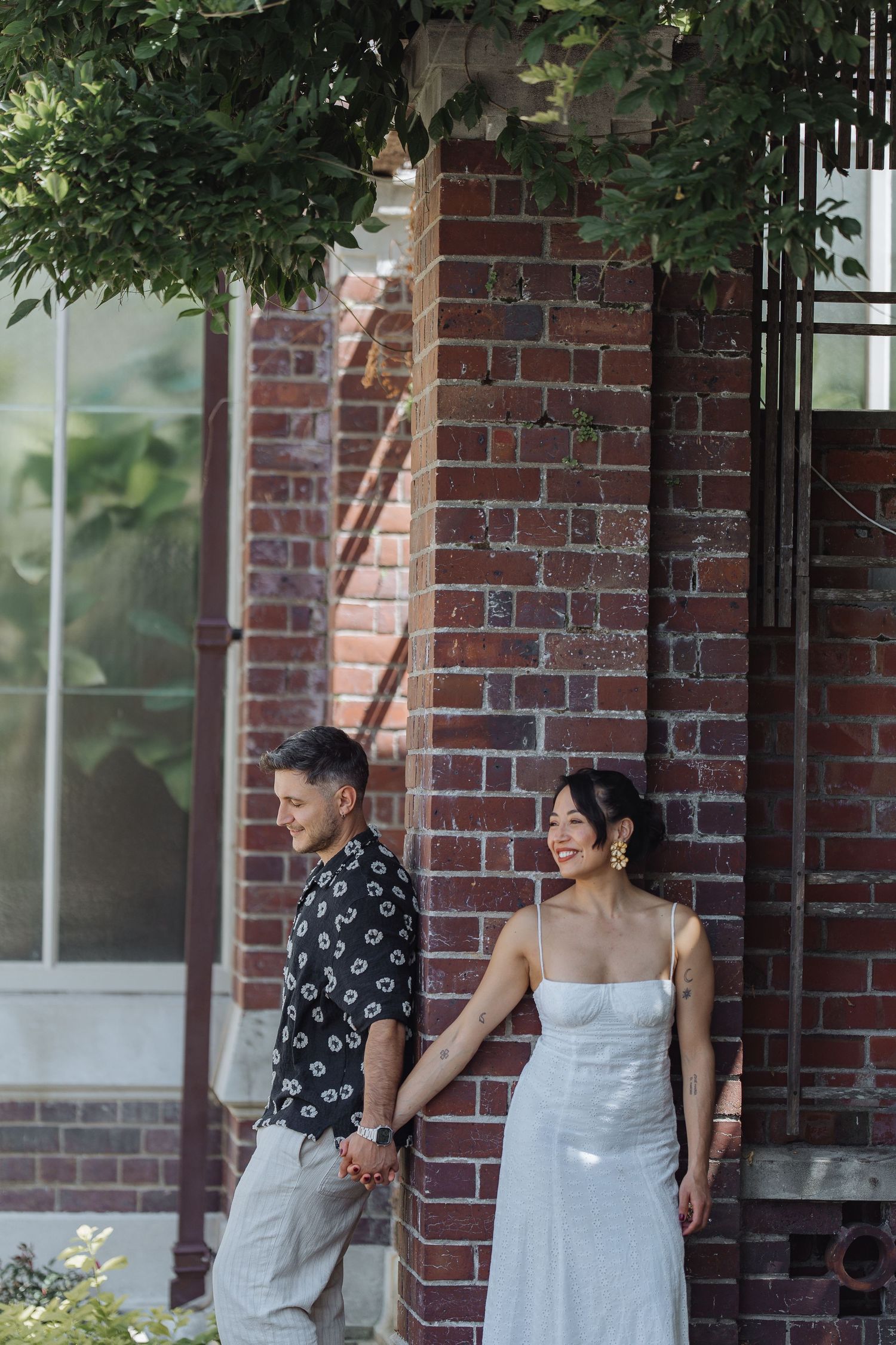 Romantic couple sharing a laugh while standing by a brick building with lush foliage.