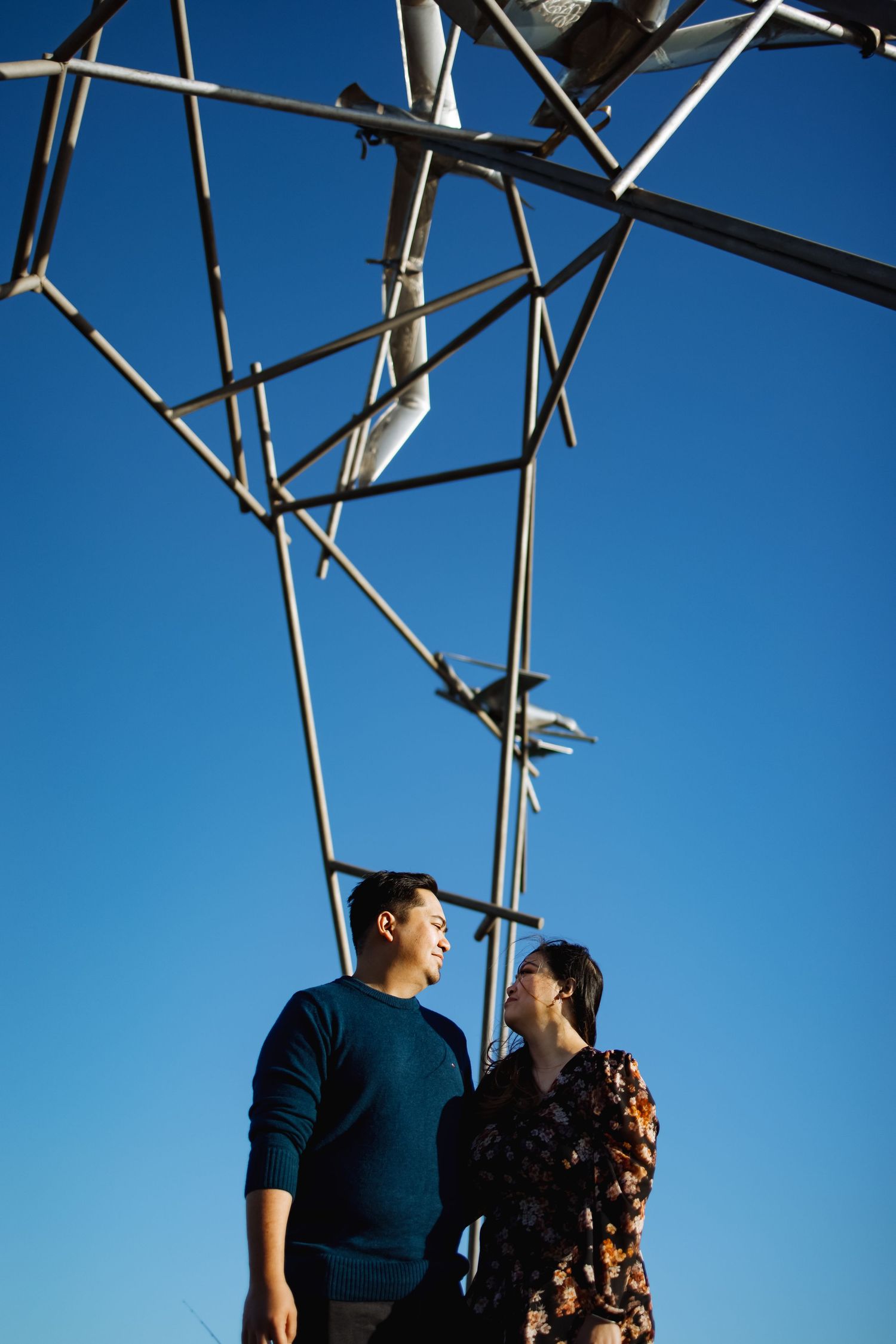 A couple stands beneath geometric metal architecture against a bright blue sky.