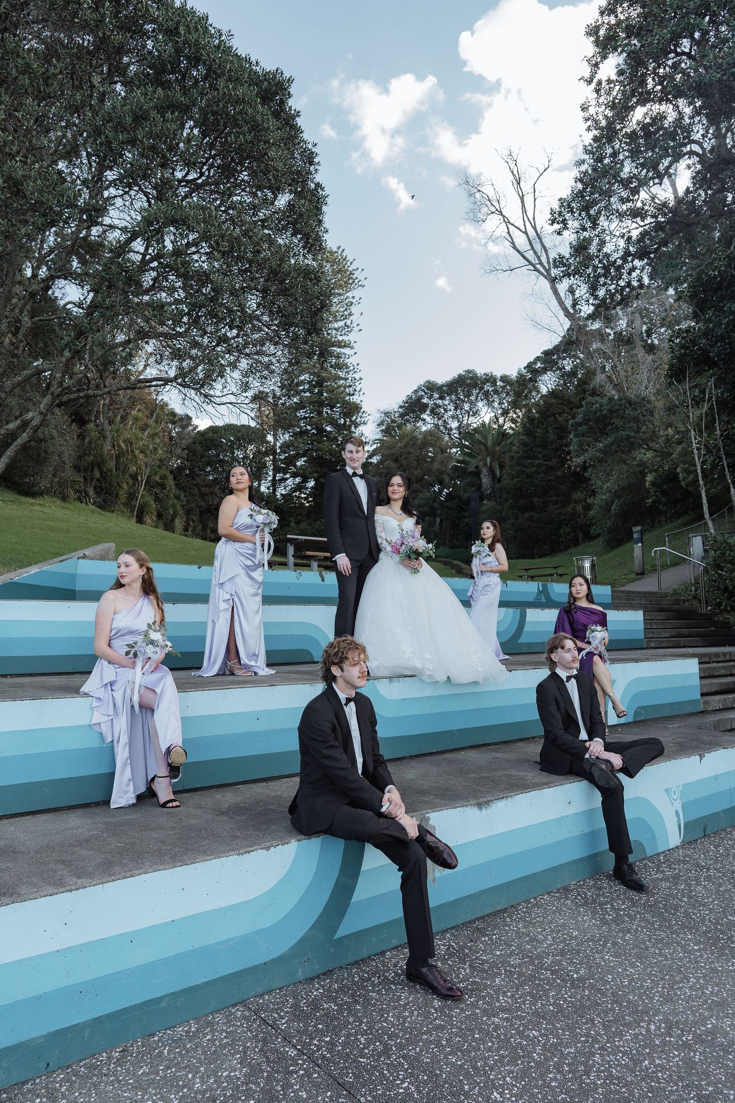Wedding party seated on blue painted concrete steps outdoors with trees in the background.