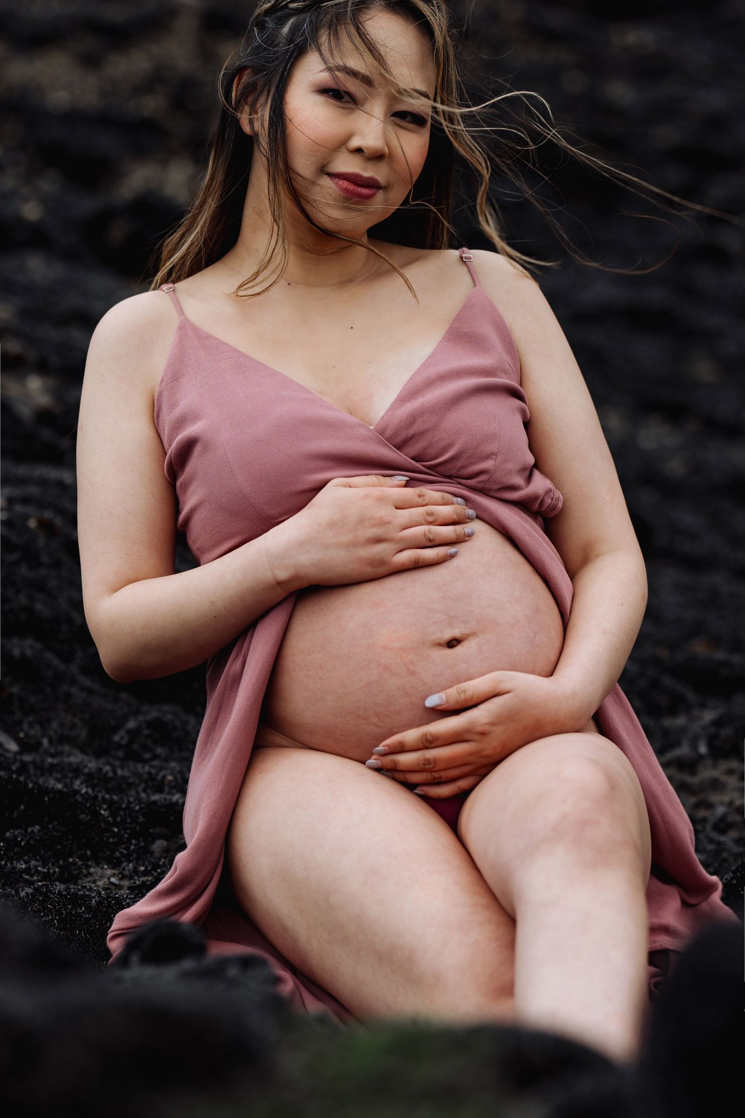 An expectant mother in a dusty rose dress poses tenderly on dark rocks.