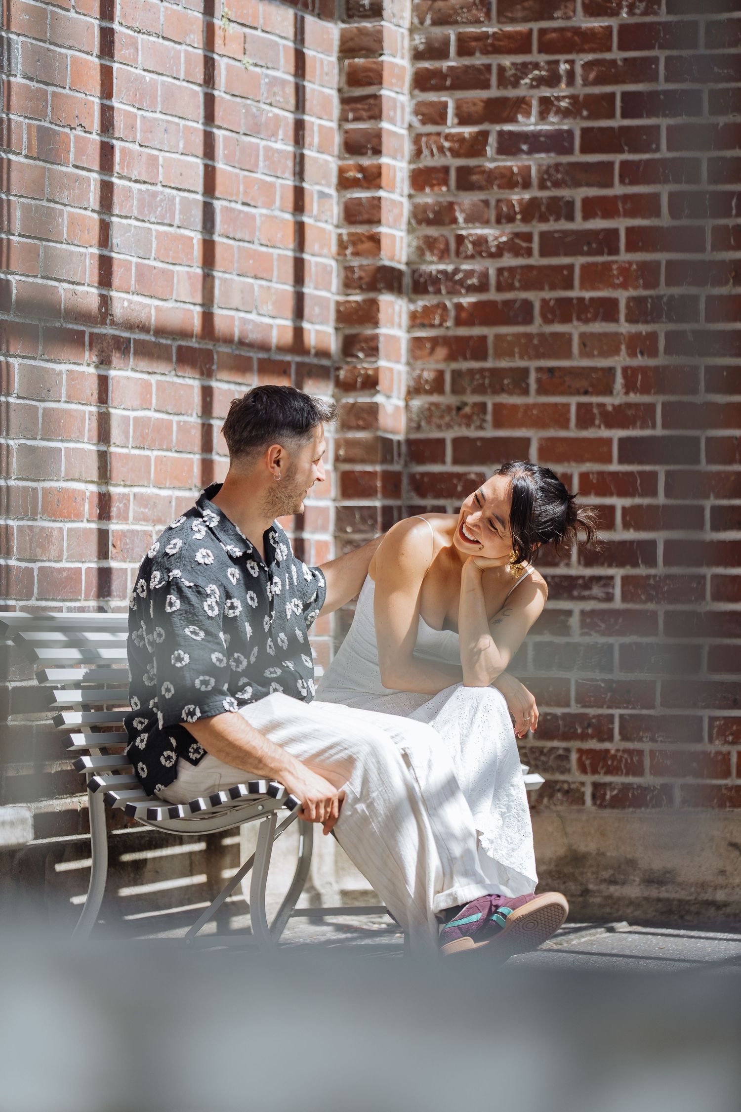 A couple relaxes on a bench against a brick wall backdrop in natural lighting.