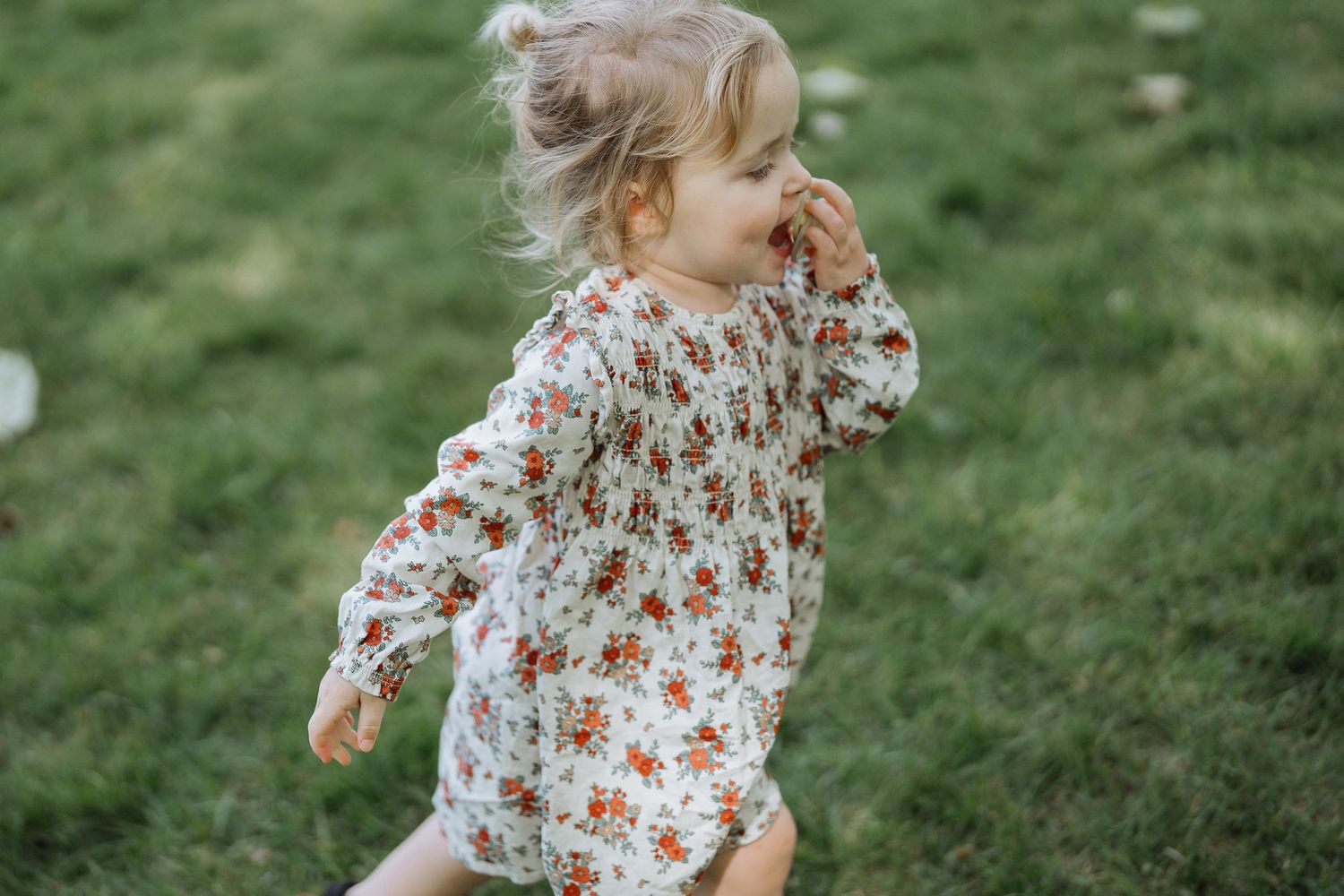 A toddler in a floral dress runs playfully through green grass on a sunny day.