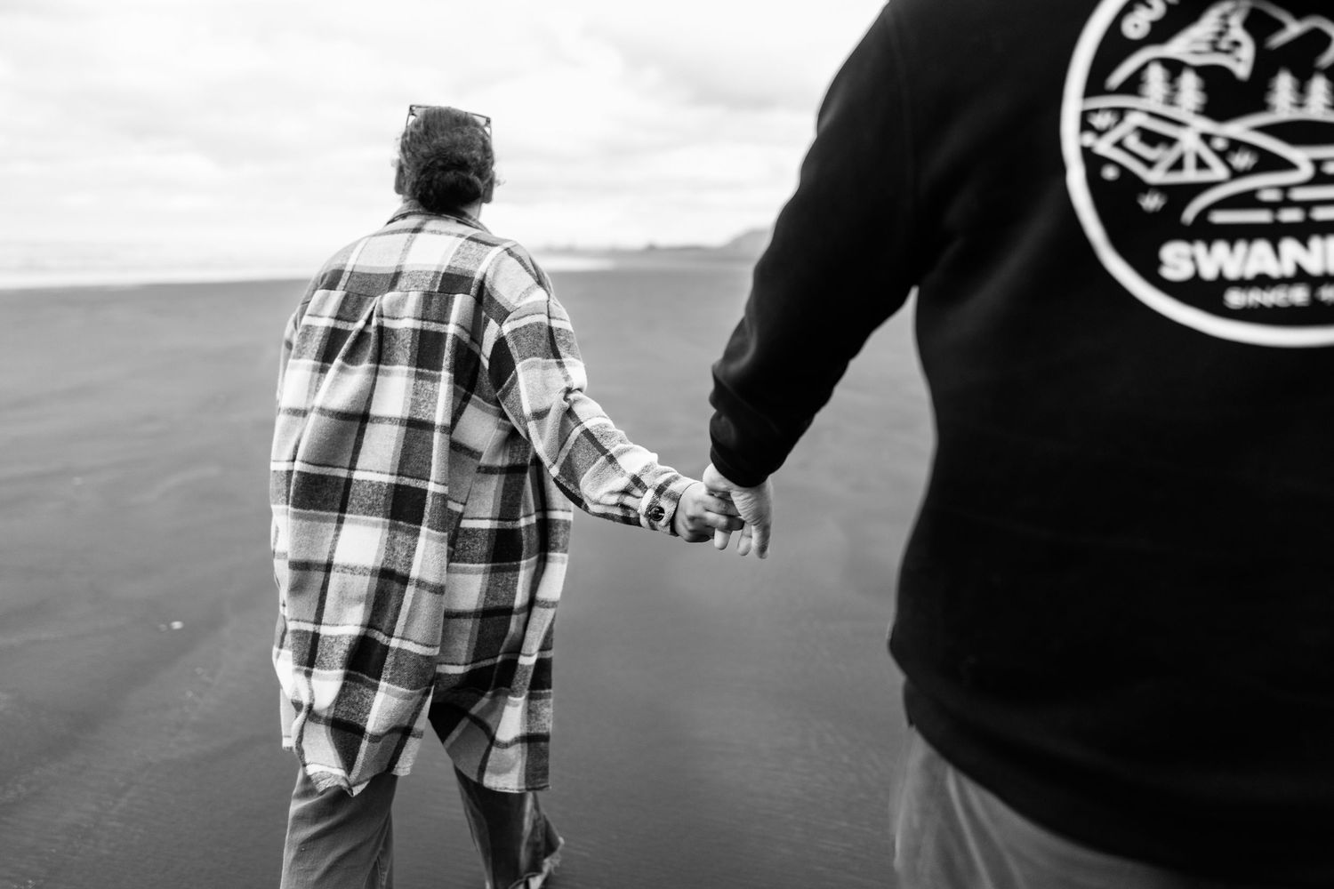 Black and white photo of two people holding hands on a beach, one wearing a plaid shirt and the other a Swans sweatshirt.