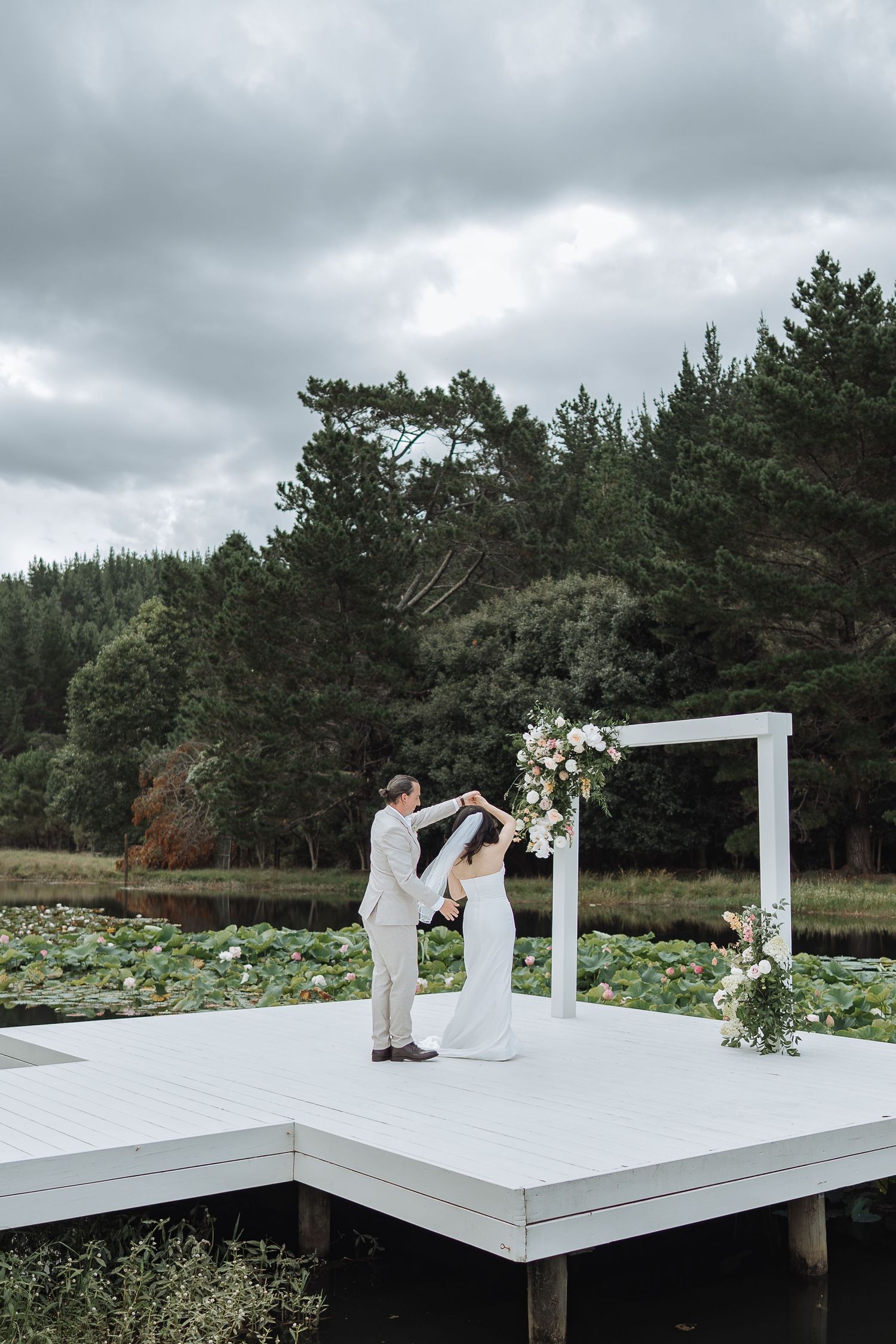 Modern minimalist wedding ceremony on white platform with floral arch and pine forest backdrop.