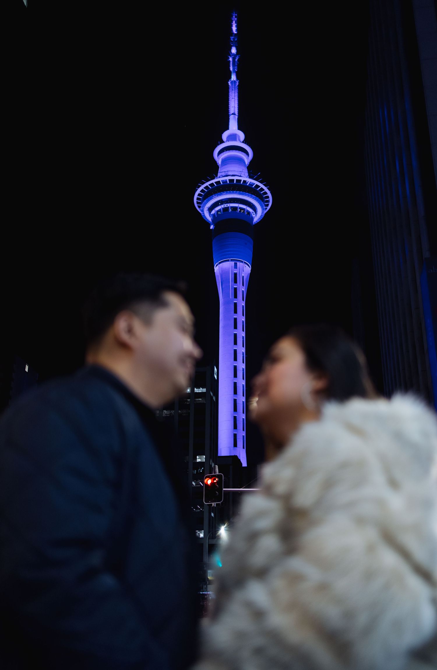 A couple stands against the illuminated blue Sky Tower in Auckland at night.