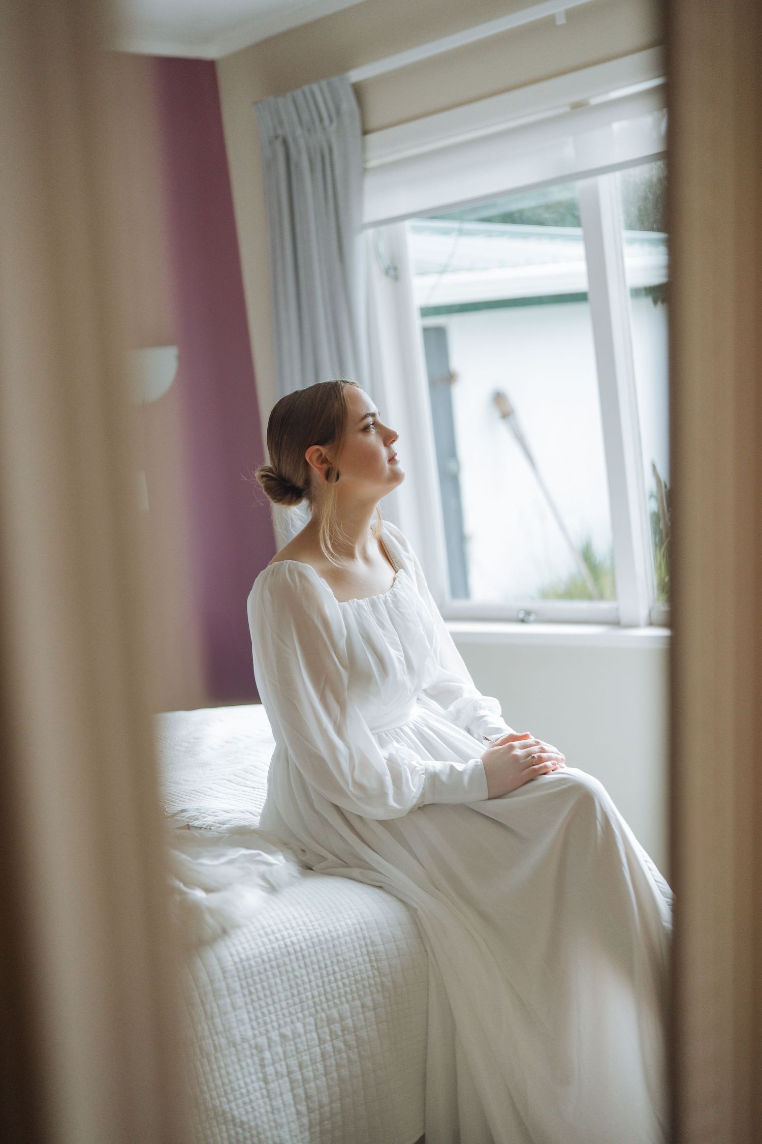 A bride in a vintage-style white dress sits thoughtfully by a window in natural light.