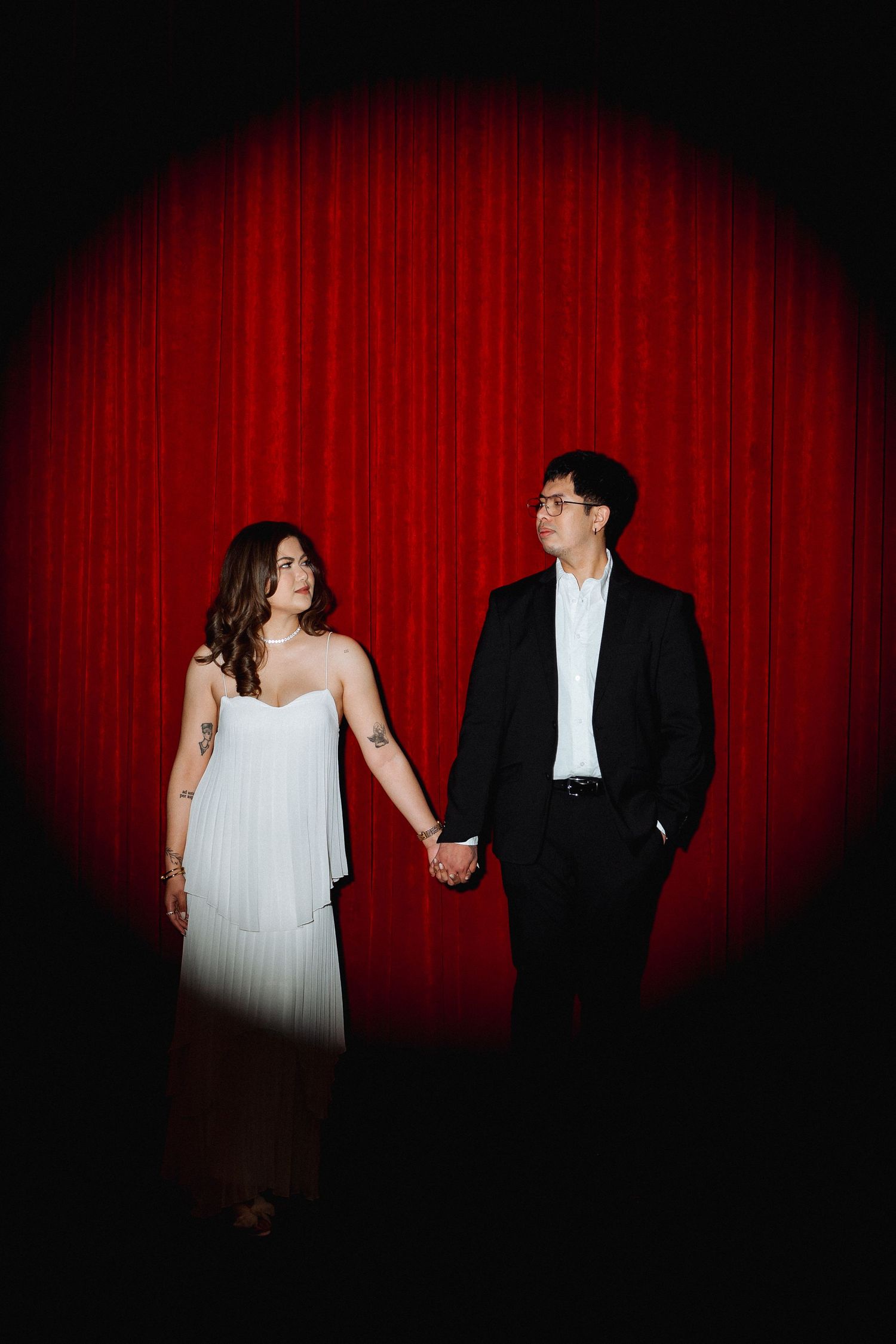 A couple in formal attire poses against a red curtain backdrop in dramatic spotlight lighting.