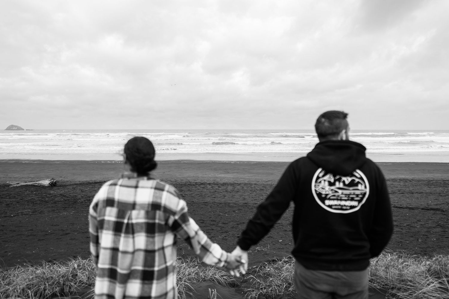 Couple holding hands facing ocean beach in black and white photo with dramatic cloudy sky.