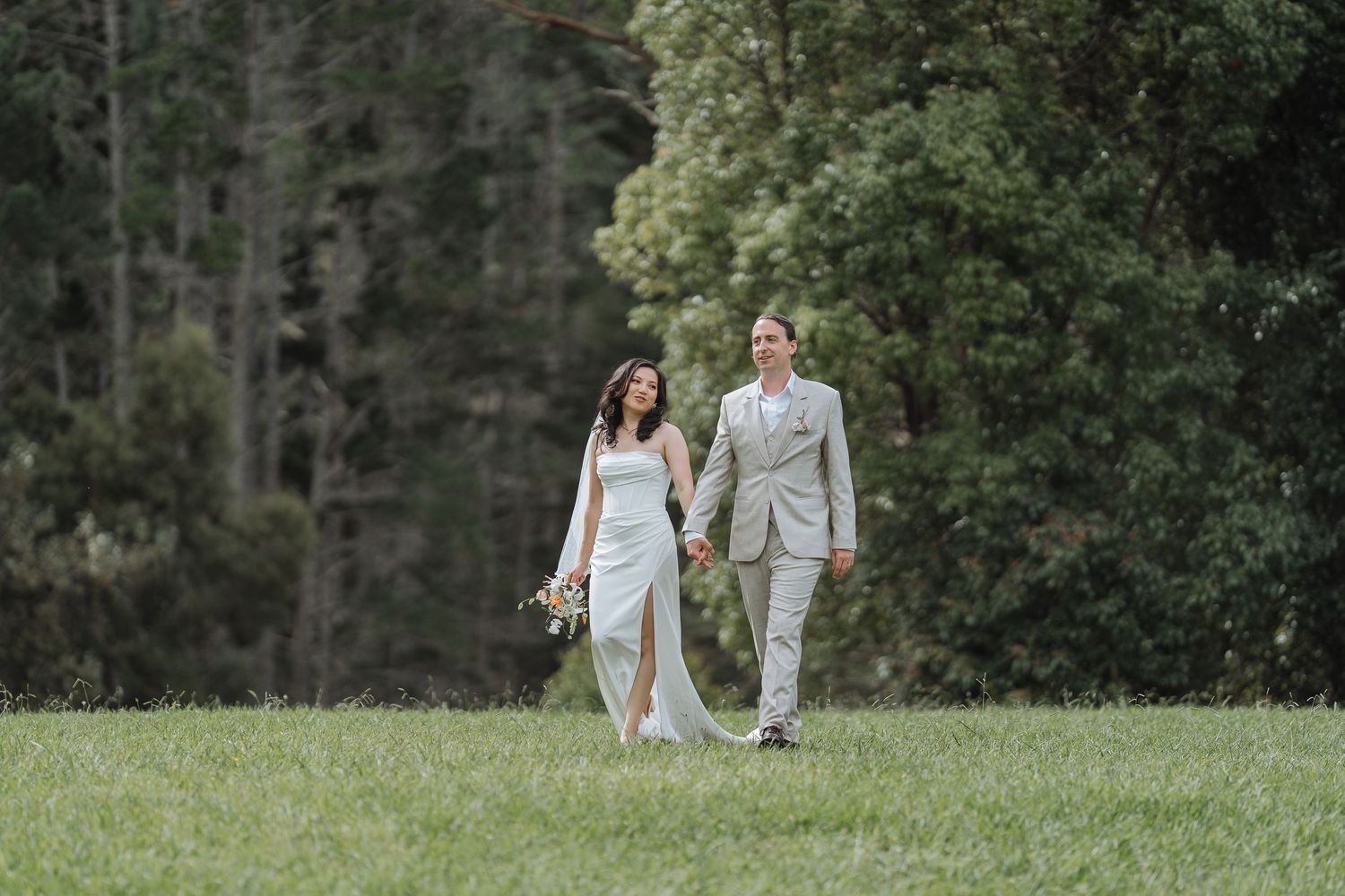 Wedding couple walking hand in hand across a grassy field with forest backdrop.