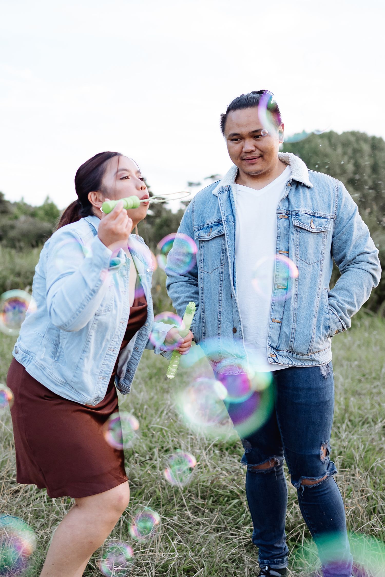 Playful couple blowing bubbles together in a grassy field wearing matching denim jackets.