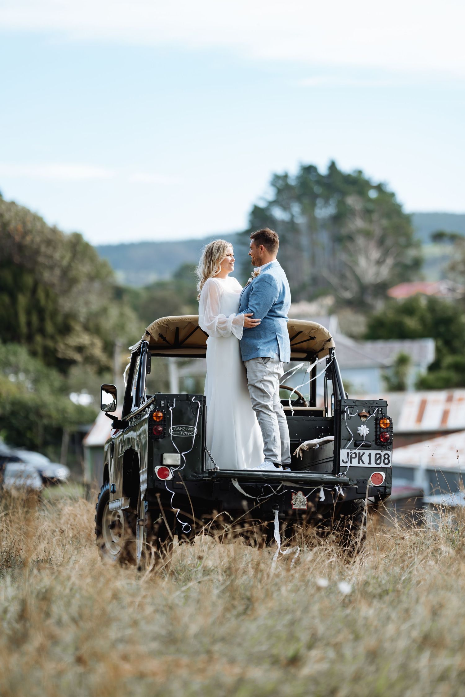 Couple sitting together on vintage car in grassy field during sunset.