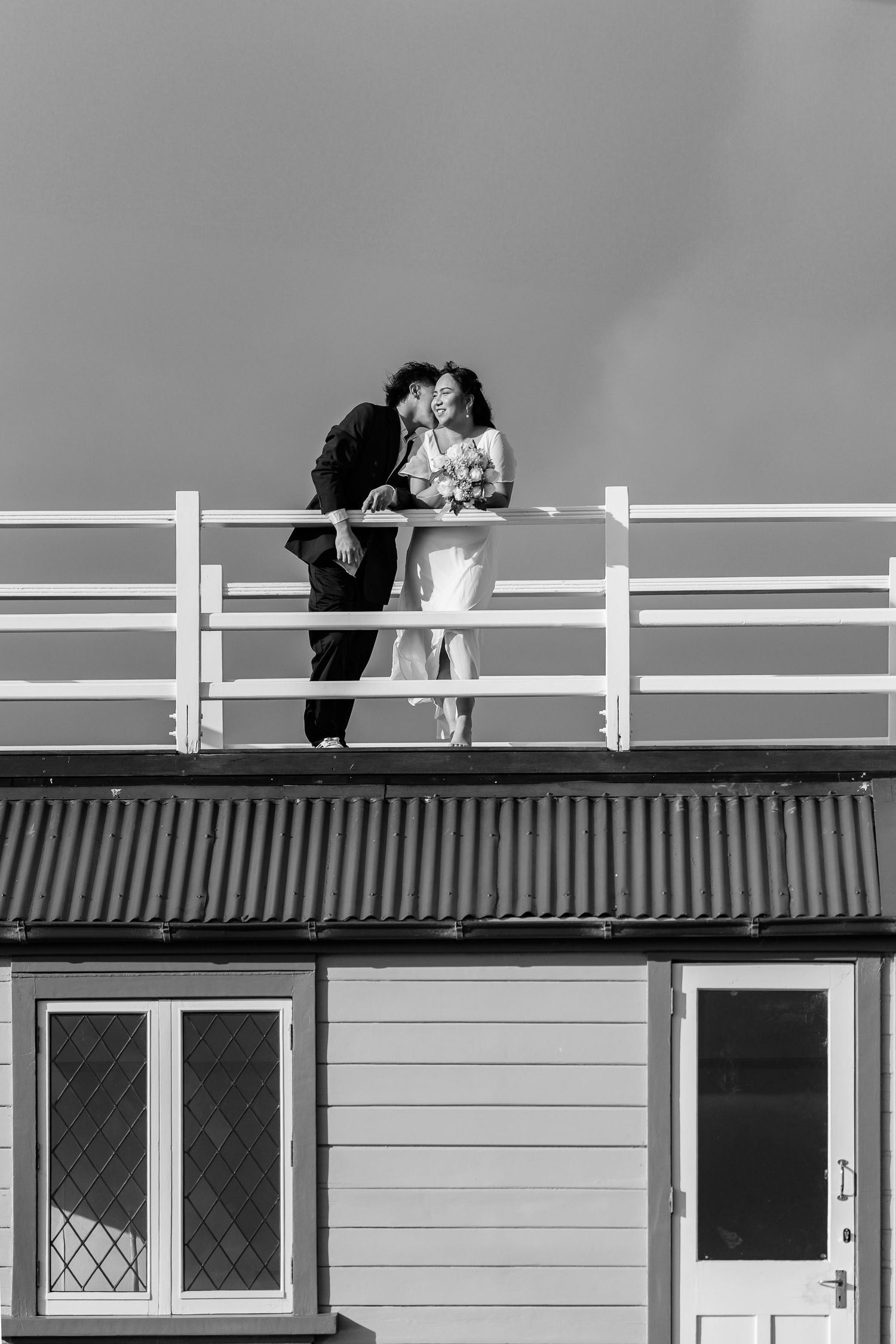 A couple embraces on a white railing against a suburban building backdrop in black and white.