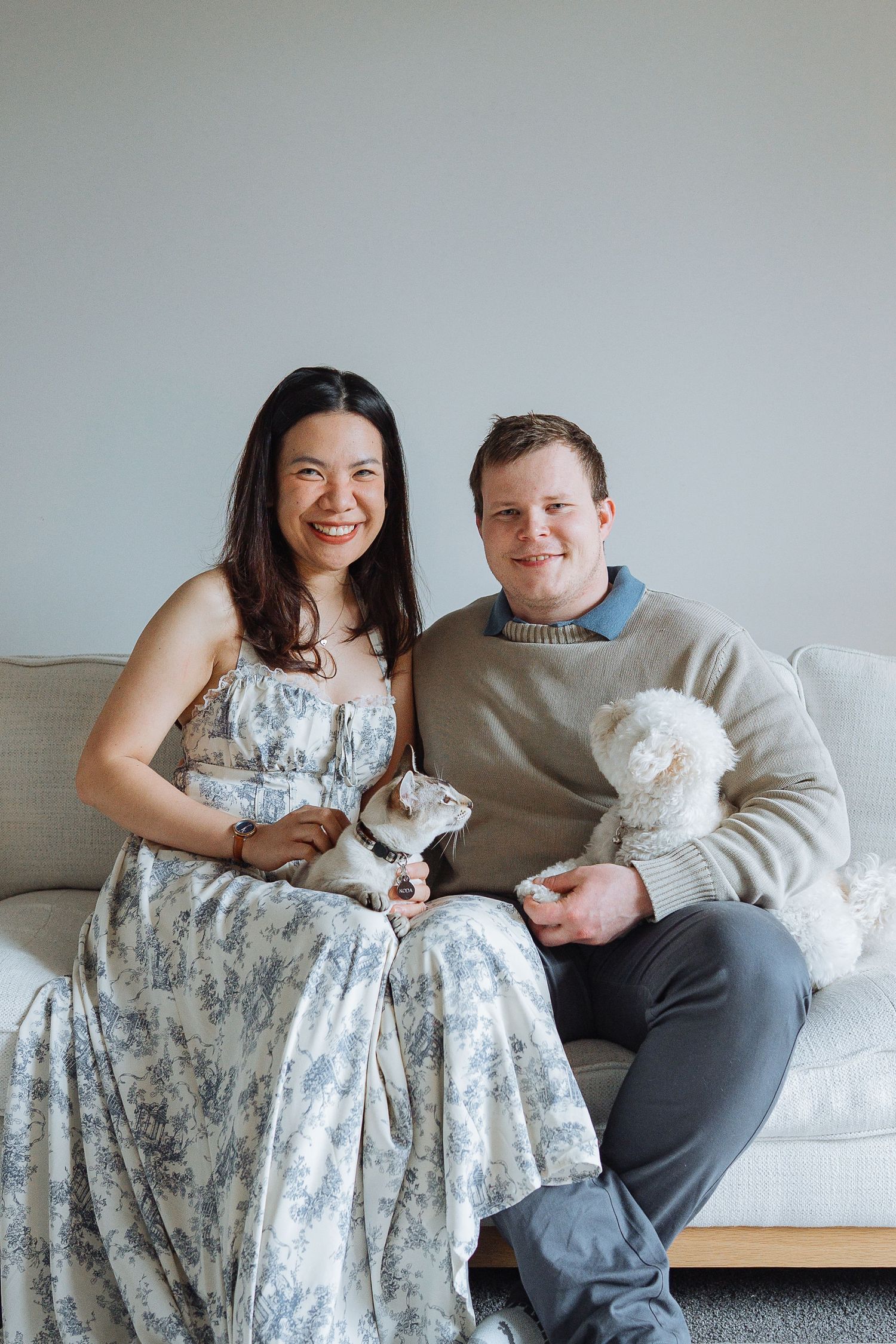 A couple poses on a couch with small white dogs in a bright indoor setting.