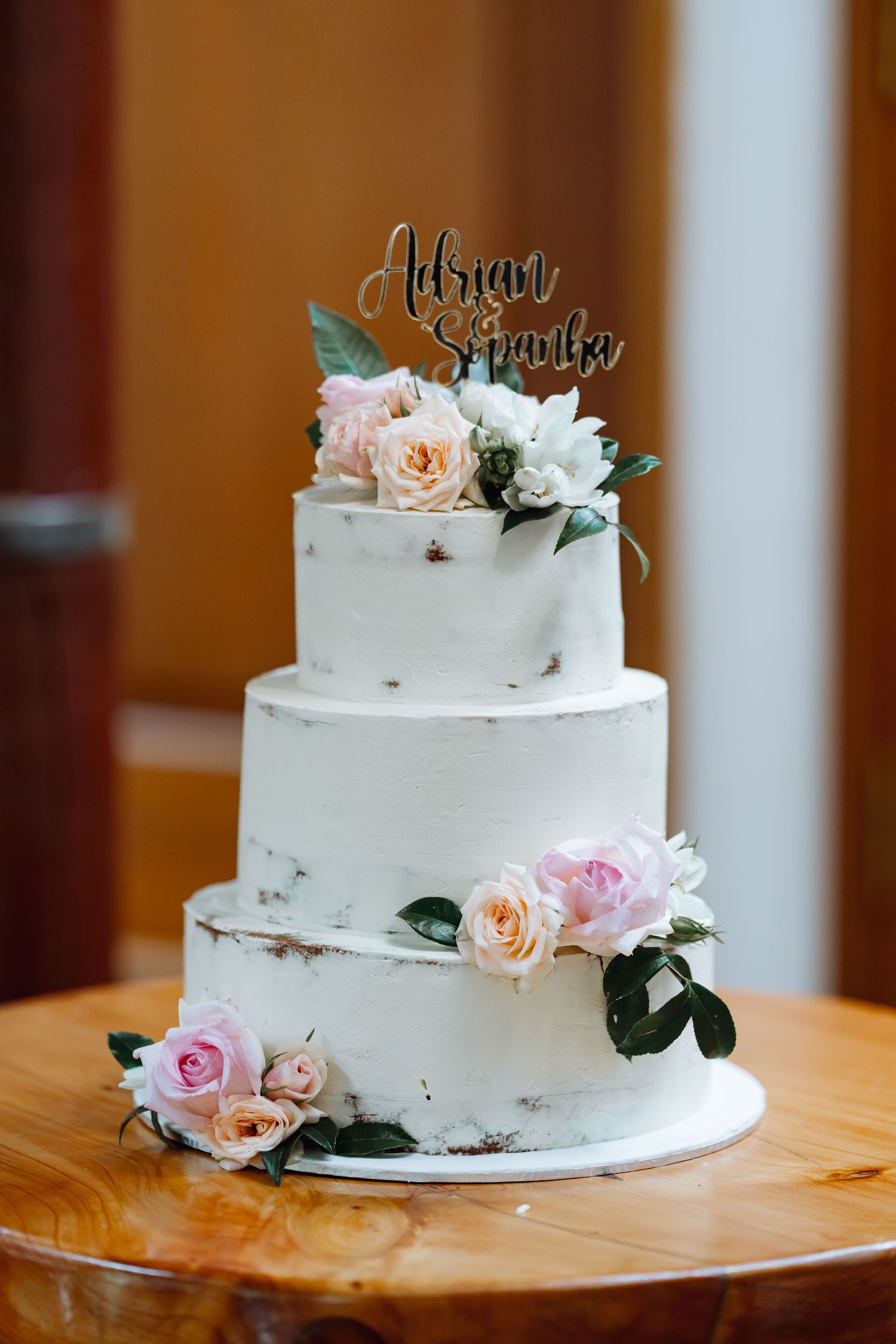 Three-tiered white semi-naked wedding cake decorated with fresh pink roses and a custom name topper.