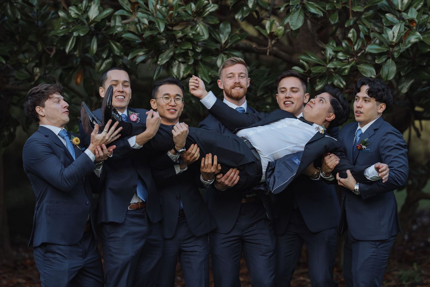 Groomsmen in navy suits pose playfully in a garden setting during a wedding photoshoot.